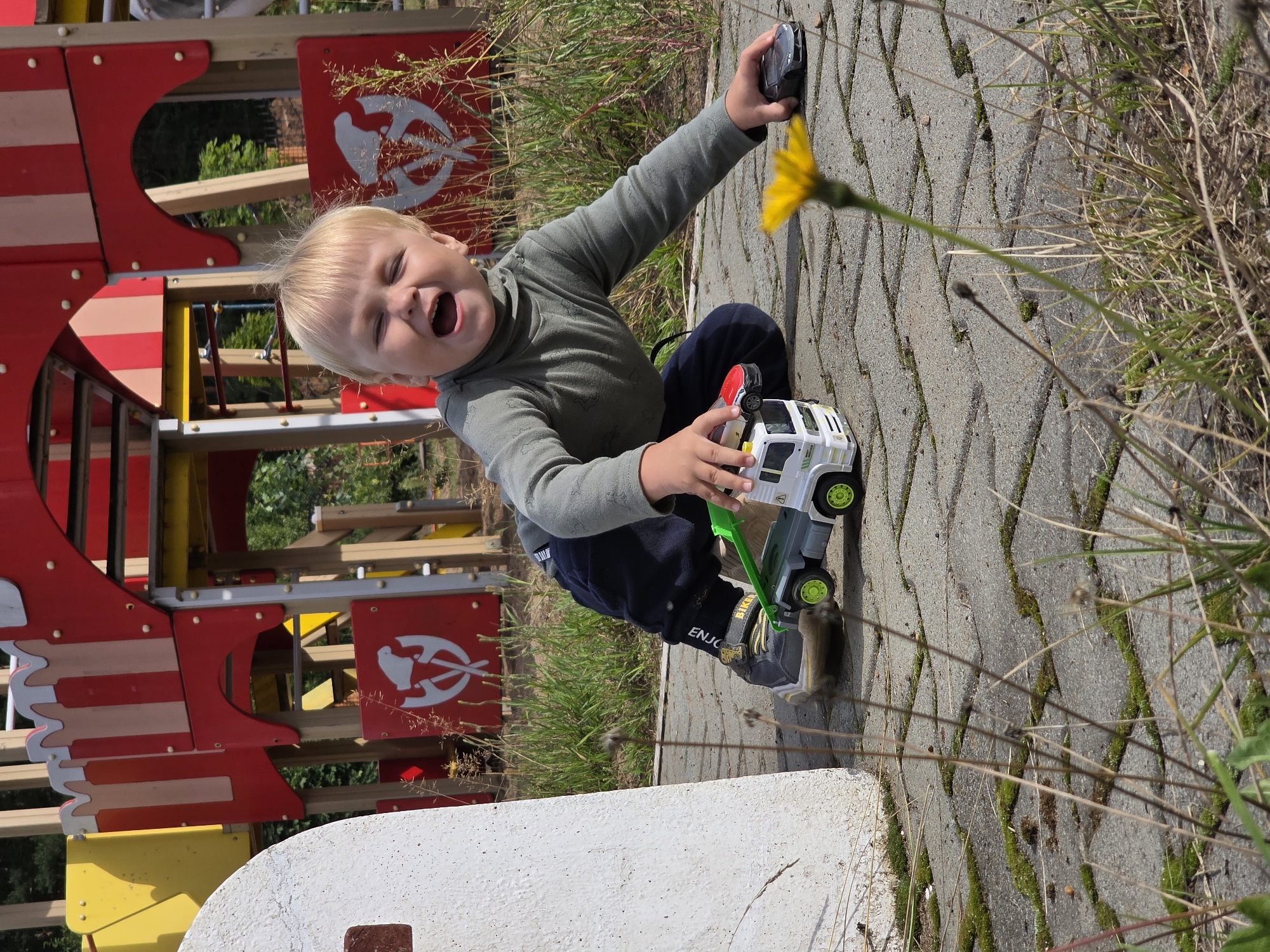 A blonde-haired child wearing a green turtleneck and dark pants kneels on a grey paved surface covered with moss and dry grass. The child is playing with toy vehicles, specifically a white and green tow truck with a red car on its flatbed, and a dark grey car. The child's mouth is open in a joyous expression. In the background, a red and yellow playground structure with a fortress-like design is visible. The location is a playground in Chaikovsky, Russia. A single yellow flower is in the foreground, slightly out of focus.