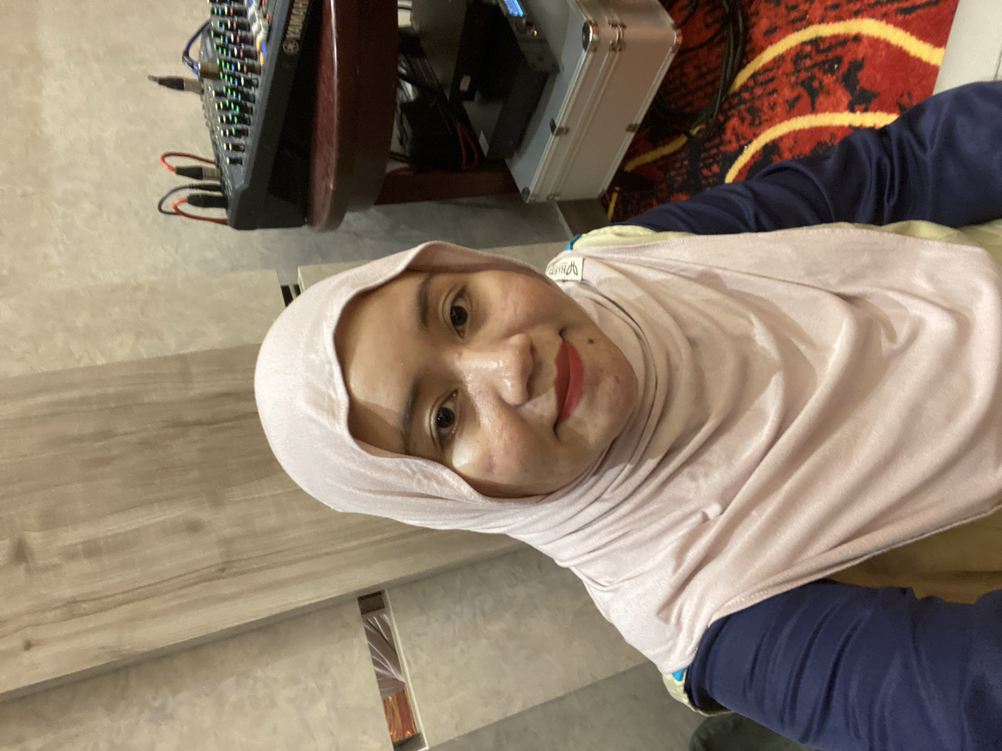 A young woman wearing a light pink hijab and a navy blue shirt poses for a selfie. She has makeup on her face, including lipstick. Behind her, to the right, is a wooden table with a sound mixer and some electronic equipment. The background includes a wall with a wood-like texture and a patterned carpet on the floor. The image appears to have been taken indoors, possibly in Banjarbaru, Indonesia, given the context of the request.