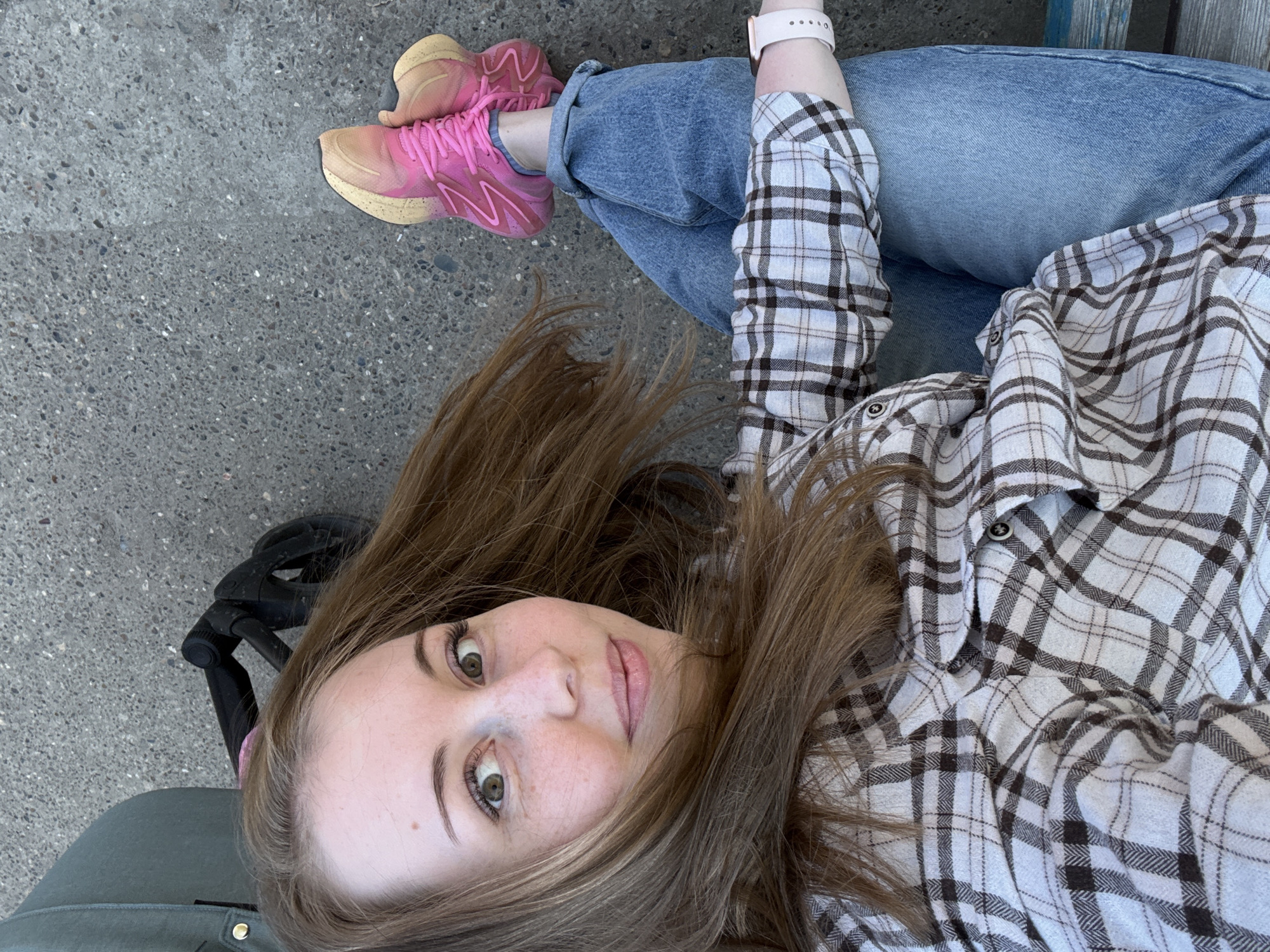 A medium shot captures a fair-skinned individual with light brown hair and green eyes. The person is wearing a white and brown plaid shirt over light blue jeans. A white smartwatch is visible on their left wrist. They are seated on a blue wooden bench, with pink sneakers on their feet. A black stroller is partially visible to the left. The background is a gray concrete surface. The location is the city of Unknown, Unknown.