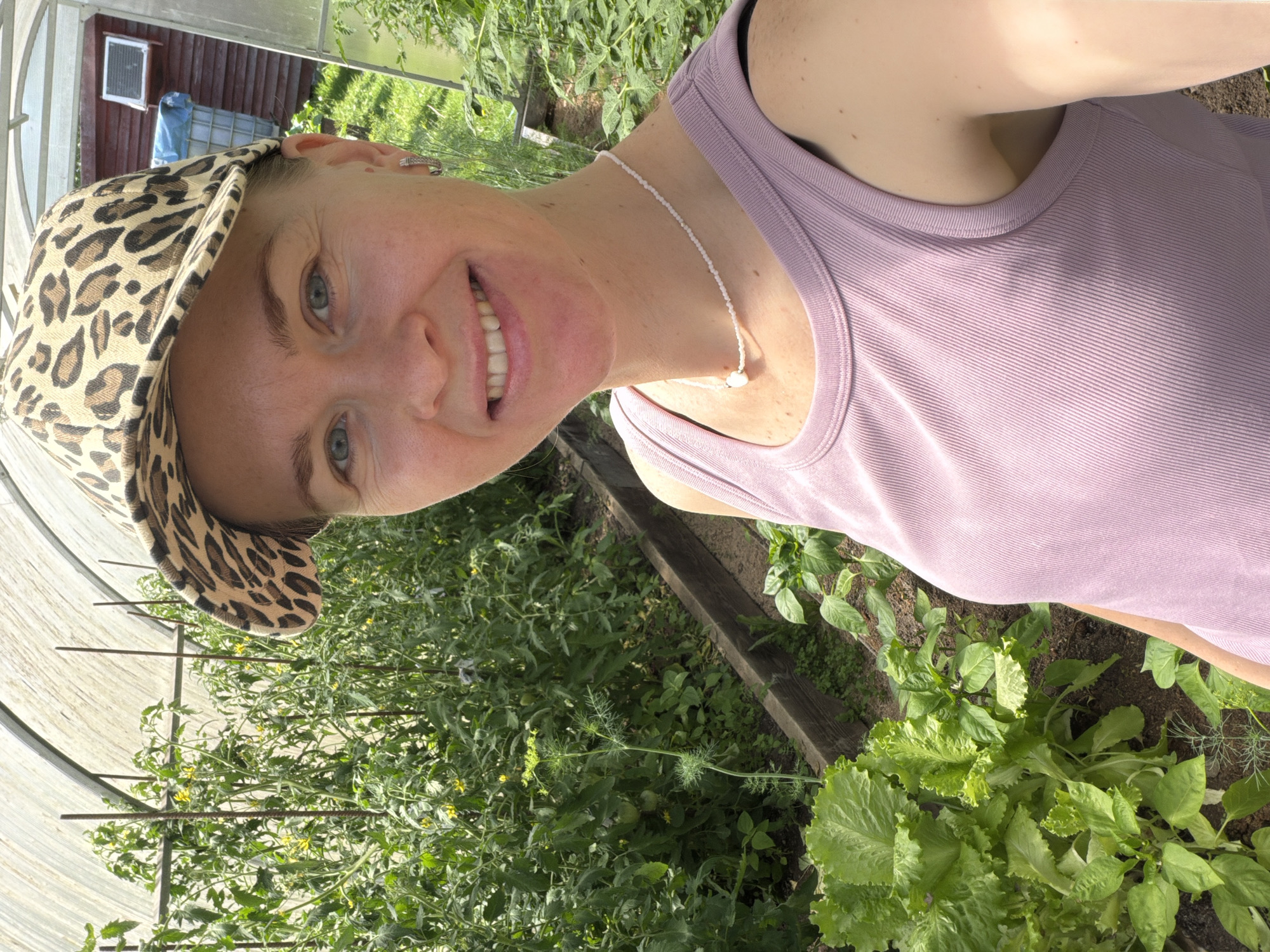 A woman with light skin and blue eyes, wearing a leopard-print baseball cap and a pink ribbed tank top, is taking a selfie in a greenhouse. She is smiling and has a pearl necklace with a white pendant. To her left, a row of tomato plants with yellow flowers grows in a raised garden bed. In the foreground to the left, a large head of lettuce is visible, and to the right, smaller pepper plants are growing. The greenhouse has a white translucent roof and wooden sides. In the background, there is a red building with a window. The scene appears to be outdoors in Khekhtsir, Russia, under a sunny sky.