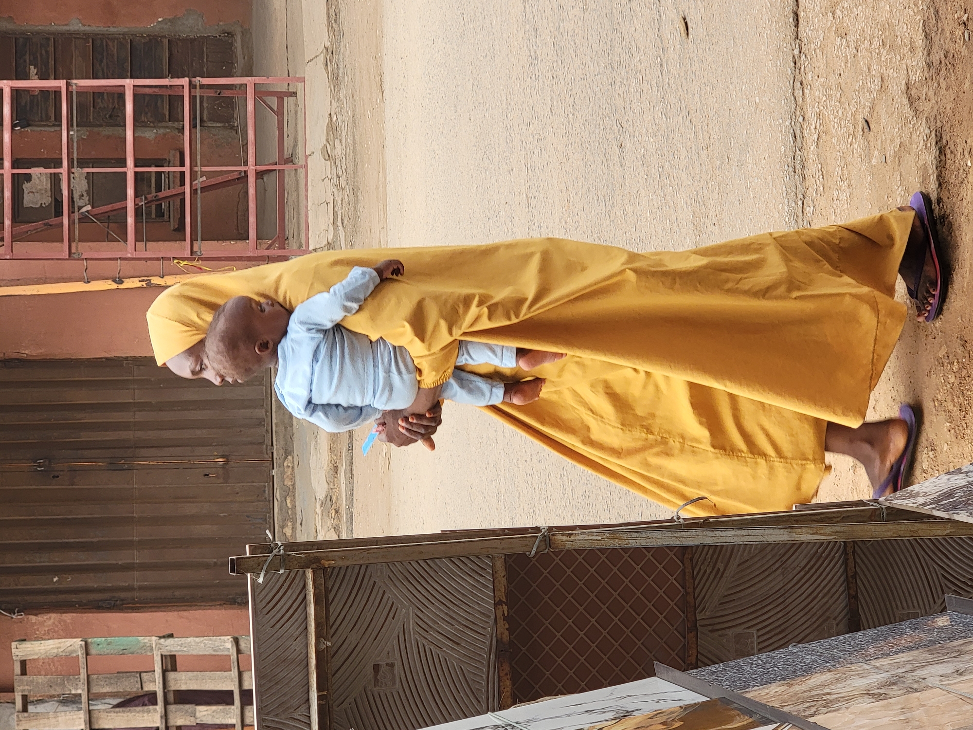 This image, captured in Maiburiji, Nigeria, depicts an adult carrying a baby outdoors on a sunny day. The adult, wearing a long, flowing yellow garment, possibly an abaya or dress, and a matching head covering, holds the baby on their left side. They are also wearing purple sandals. The baby is dressed in a light blue long-sleeved top and light blue trousers, with its head turned towards the adult's shoulder. The adult looks down, appearing calm and attentive to the baby, who is holding a small blue object in its right hand.
The setting appears to be a dusty or unpaved street or path. In the background, there are various buildings and structures. A weathered reddish-brown building with a metal grate or railing is visible, along with a lighter, textured wall. Towards the lower left, some wooden pallets are stacked. Part of a decorative metal gate with intricate patterns is also visible in the mid-ground. Pieces of patterned tiles are leaning against something in the foreground on the right. The strong daylight and visible shadows indicate a clear, bright day, likely midday or early afternoon. There is no visible text in the image.