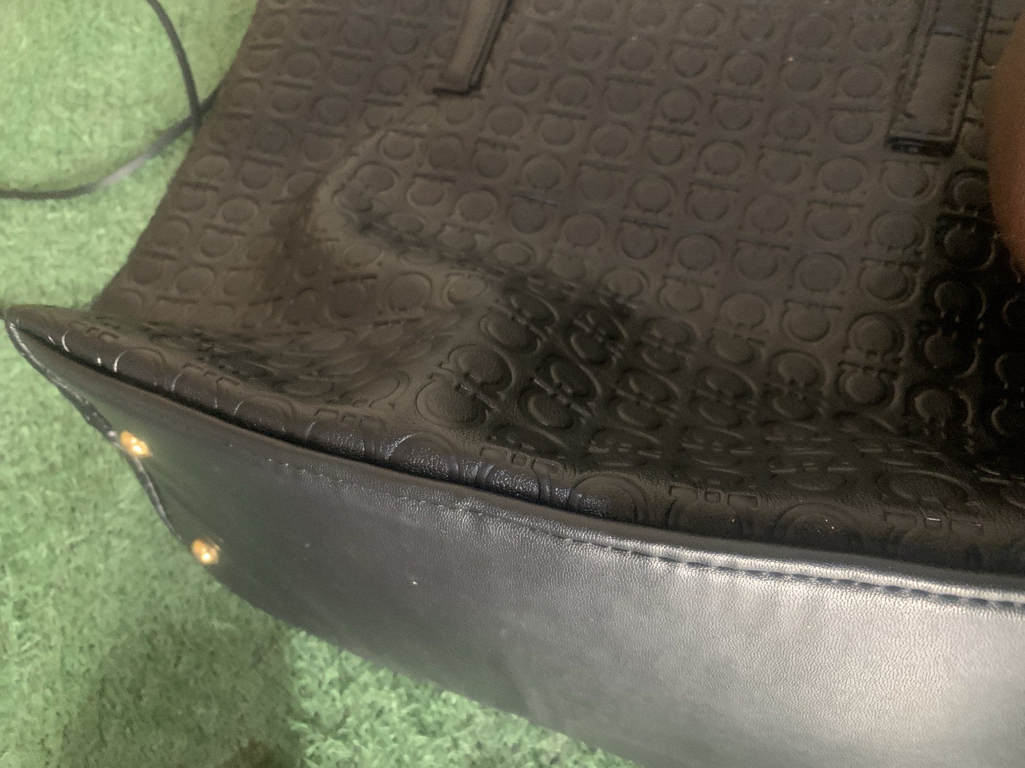 A black leather handbag with a repeating Gancini pattern is placed on a green, textured surface, possibly artificial turf. The handbag has a smooth, black leather side panel with visible stitching. Three small, golden studs are attached to the edge where the smooth leather meets the patterned section. The patterned section of the bag is embossed with the Gancini logo. A dark cable is visible in the upper right corner, extending out of frame. The scene appears to be indoors or in a sheltered outdoor area, with no specific urban or natural landmarks to indicate a location in Jauro Yinu, Nigeria.