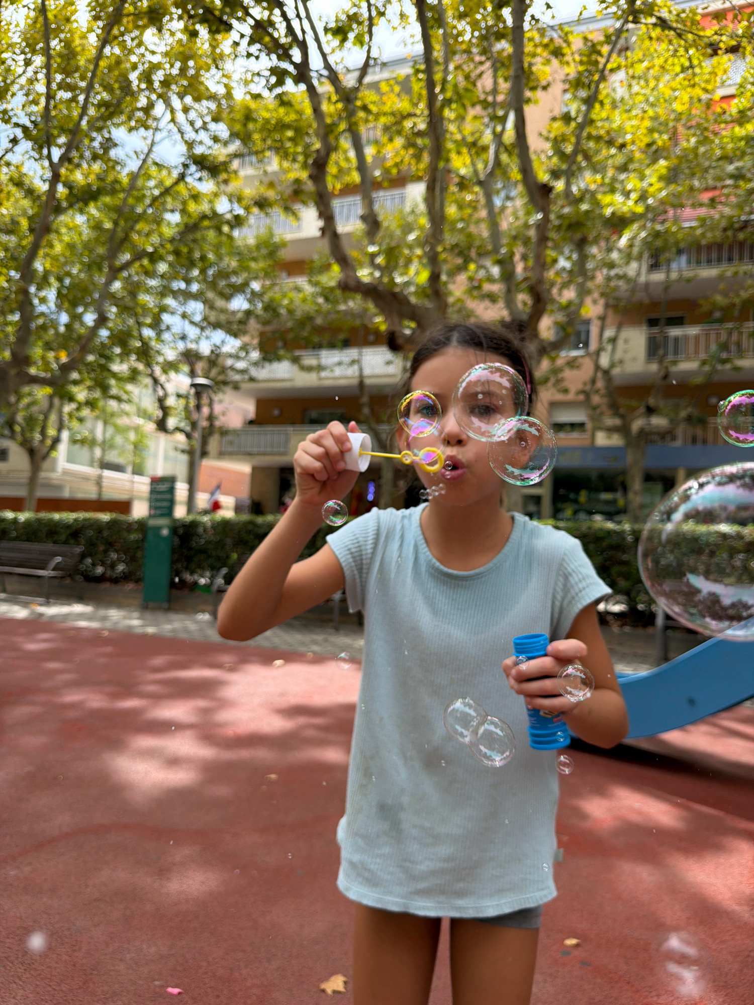 A young girl is blowing bubbles in a park in Salou, Spain. She is wearing a light blue t-shirt and gray shorts. She is holding a bubble wand in her right hand and a bottle of bubble solution in her left. Several bubbles are floating in the air around her. In the background, there are trees and buildings. The ground is red and appears to be a playground surface.