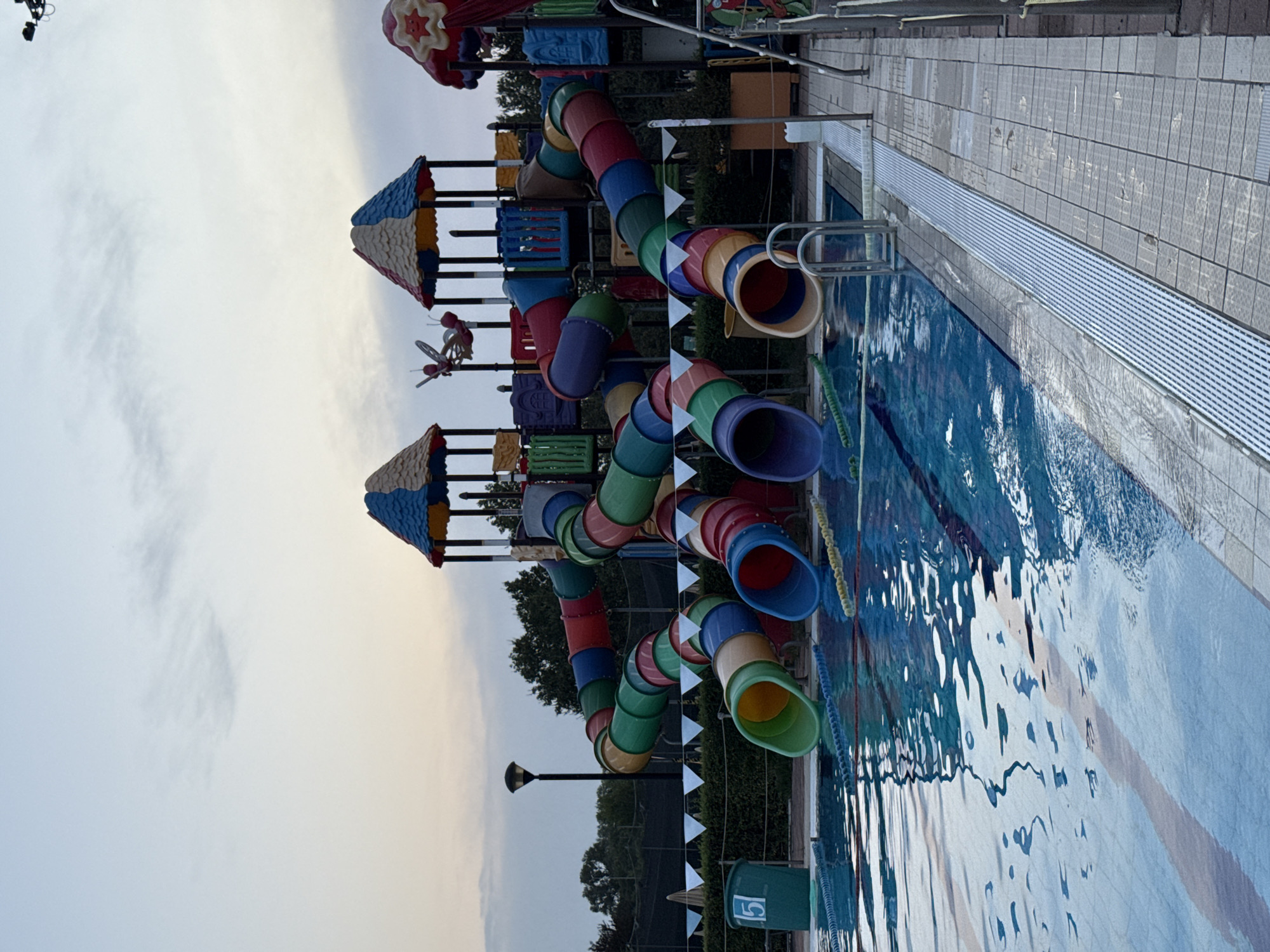 The image depicts an outdoor swimming pool area in Parma, Italy, at dusk. The foreground features a swimming pool with lane markings and a tiled edge. To the right, a metal ladder descends into the water. Beyond the pool, a large, colorful playground structure with multiple winding tube slides is visible. The slides are various shades of red, blue, green, yellow, and purple. The playground has a multi-tiered design with colorful roofs and supporting structures. In the background, trees line the perimeter of the area. A lamppost stands near the left side of the image. A green trash bin with the number "5" is placed by the pool edge. The sky is overcast with a gradient of light blue and grey hues, indicating the end of the day. There are no people visible in the image.