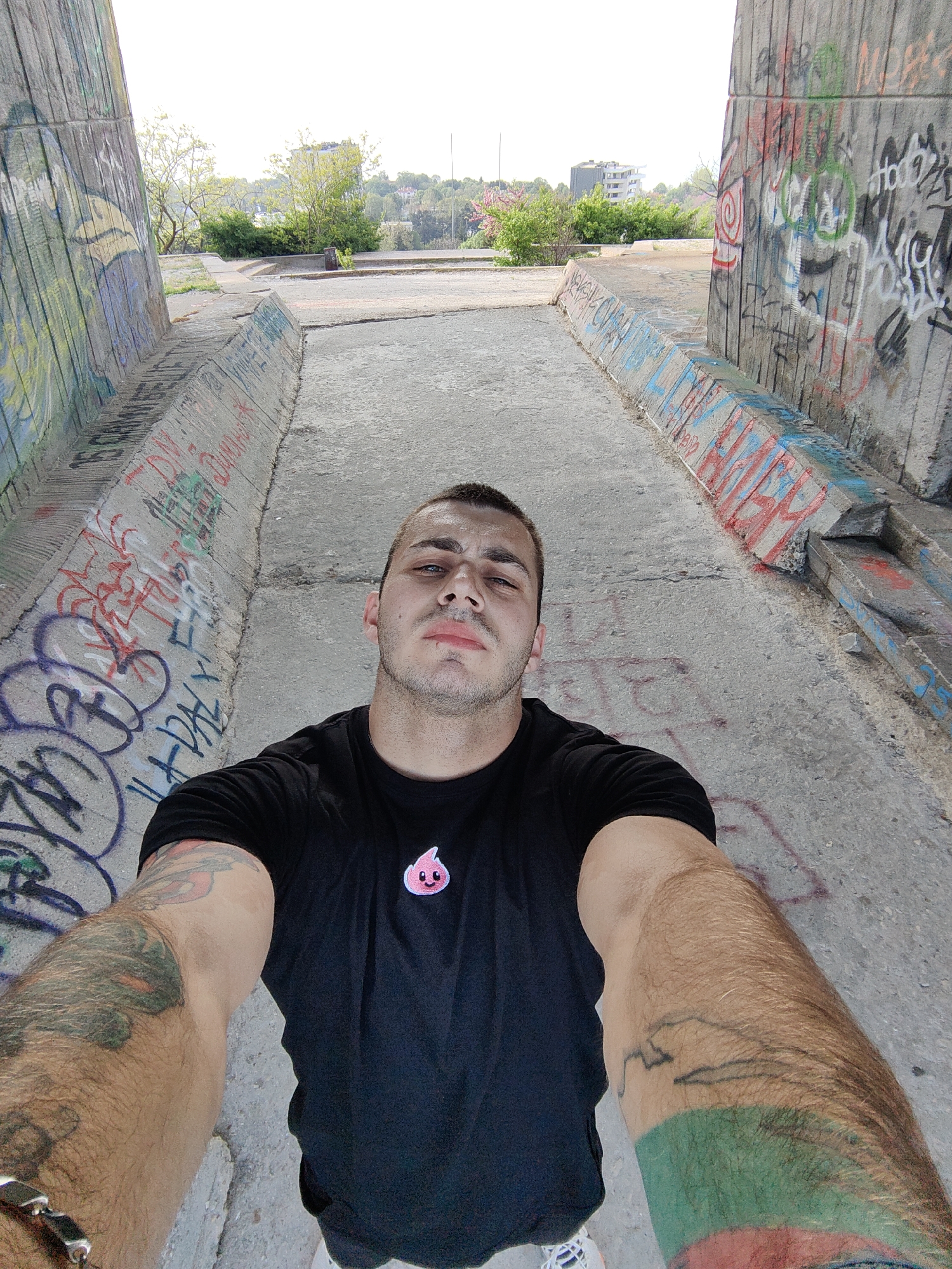 A man takes a selfie in what appears to be a pedestrian underpass or tunnel in Varna, Bulgaria, based on the urban landscape and the presence of graffiti typical of such structures. The tunnel is concrete, heavily vandalized with various graffiti tags and artworks. The man has short, dark hair and is wearing a black T-shirt with a small pink logo on the chest. His arms, which are extended toward the camera, have visible tattoos. Outside the tunnel, trees and buildings are visible, suggesting an urban environment.