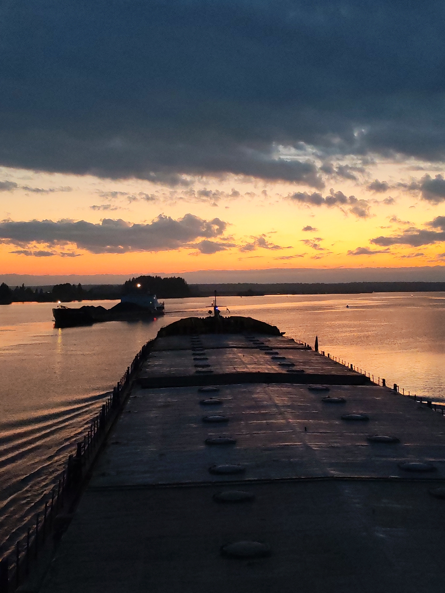 The image depicts a view from the deck of a large cargo ship at sunset. The ship's deck is visible in the foreground, featuring a flat, grey surface with circular hatches. A metal railing lines the edge of the deck. In the middle distance, the river or waterway reflects the warm colors of the setting sun. Another ship, likely a tugboat pulling a barge, is visible to the left. The background shows a distant shoreline with trees. The sky is filled with dark clouds above and illuminated by a yellow and orange glow near the horizon. The location is Unknown, Unknown.
