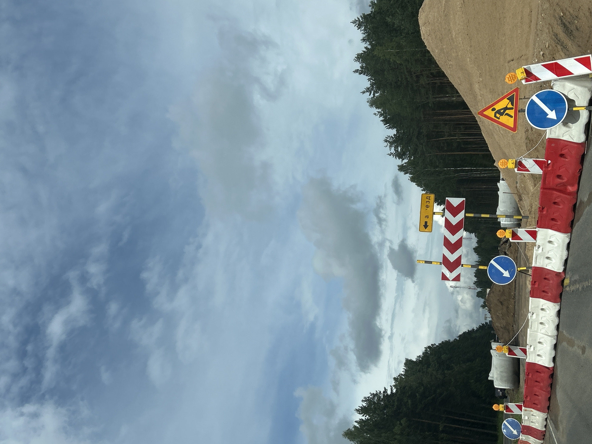 The image captures a section of road under construction, indicated by traffic barriers, signage, and a large pile of sand. The scene is located in the Плешчаніцкі сельскі Савет, Belarus, as suggested by the road signs. The road itself appears to be wet, possibly from recent rain.
On the left side of the road, a line of red and white plastic barriers marks the construction zone. Behind these barriers, large concrete pipes are visible, suggesting underground infrastructure work. A dark-colored vehicle is partially visible behind the pipes.
Several traffic signs are strategically placed to guide vehicles. A yellow sign with a black arrow pointing left reads "а - езд" which translates to "entrance" or "driveway". A large chevron sign with red and white arrows directs traffic to the right. Two blue circular signs with white arrows indicate a mandatory turn to the right. A yellow triangular sign depicts a person working with a shovel, signifying road work ahead. Additionally, two orange flashing lights are mounted on poles next to the barriers.
The background is dominated by a dense forest of pine trees lining the road on both sides. To the right, a steep mound of sand rises behind the construction area. The sky above is partly cloudy, with a mix of blue sky and scattered gray clouds. The overall impression is one of a road closure and ongoing construction project in a semi-rural or forested area.