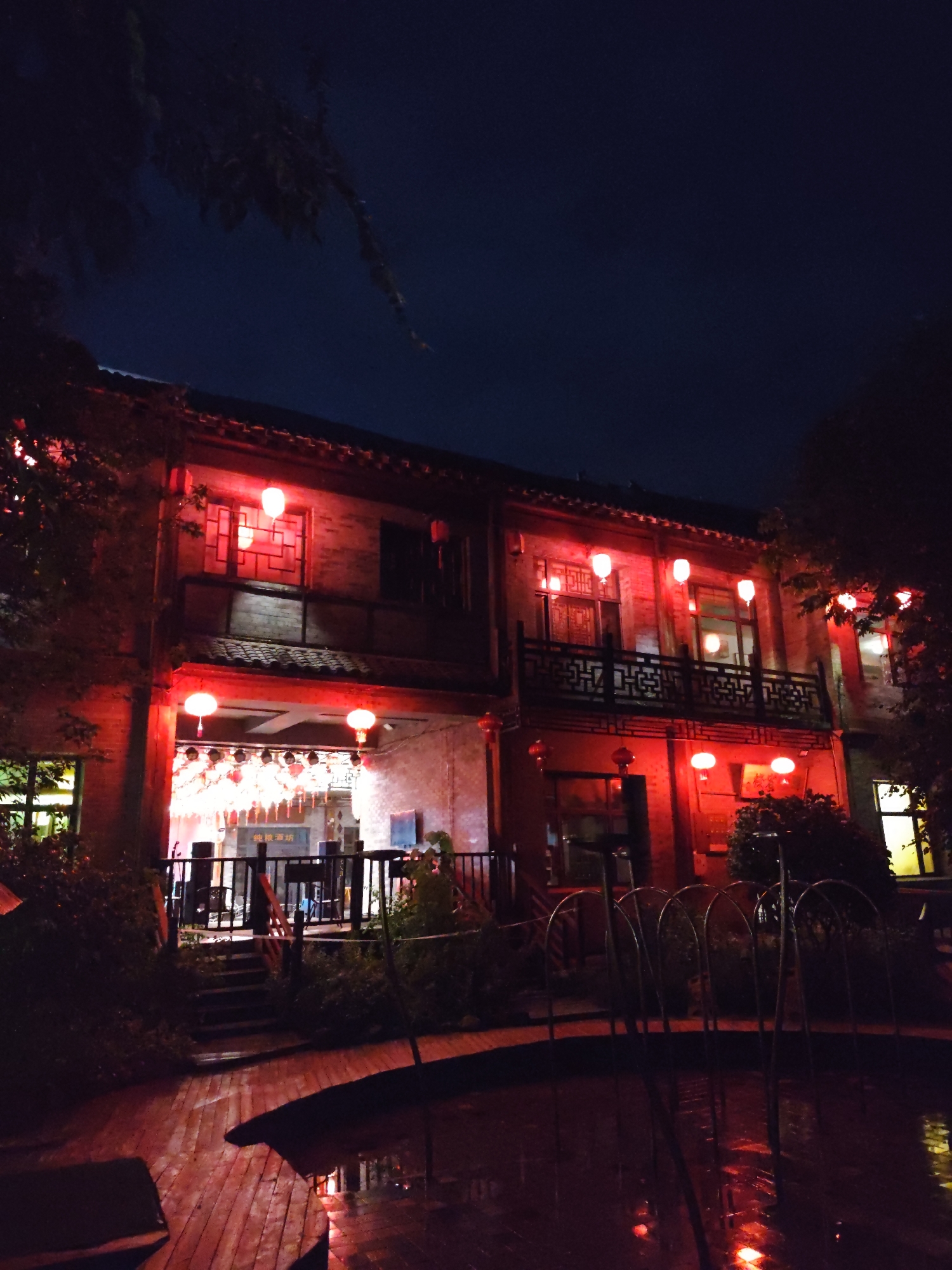 A nighttime exterior shot depicts a two-story building illuminated by red lights and traditional red lanterns. The building, with a tiled roof and brick facade, features balconies and windows. In front of the building, a stage area is visible with speakers and a backdrop. Red lanterns hang from the building and appear to be suspended above the stage. A wooden deck leads to steps ascending to the building. In the foreground, a fountain-like structure with arched metal elements is present, with its reflection visible in the water below. Trees with dark foliage frame the scene. The sky is dark blue. The location is identified as 林甸镇, China. There are no people clearly visible in the image.