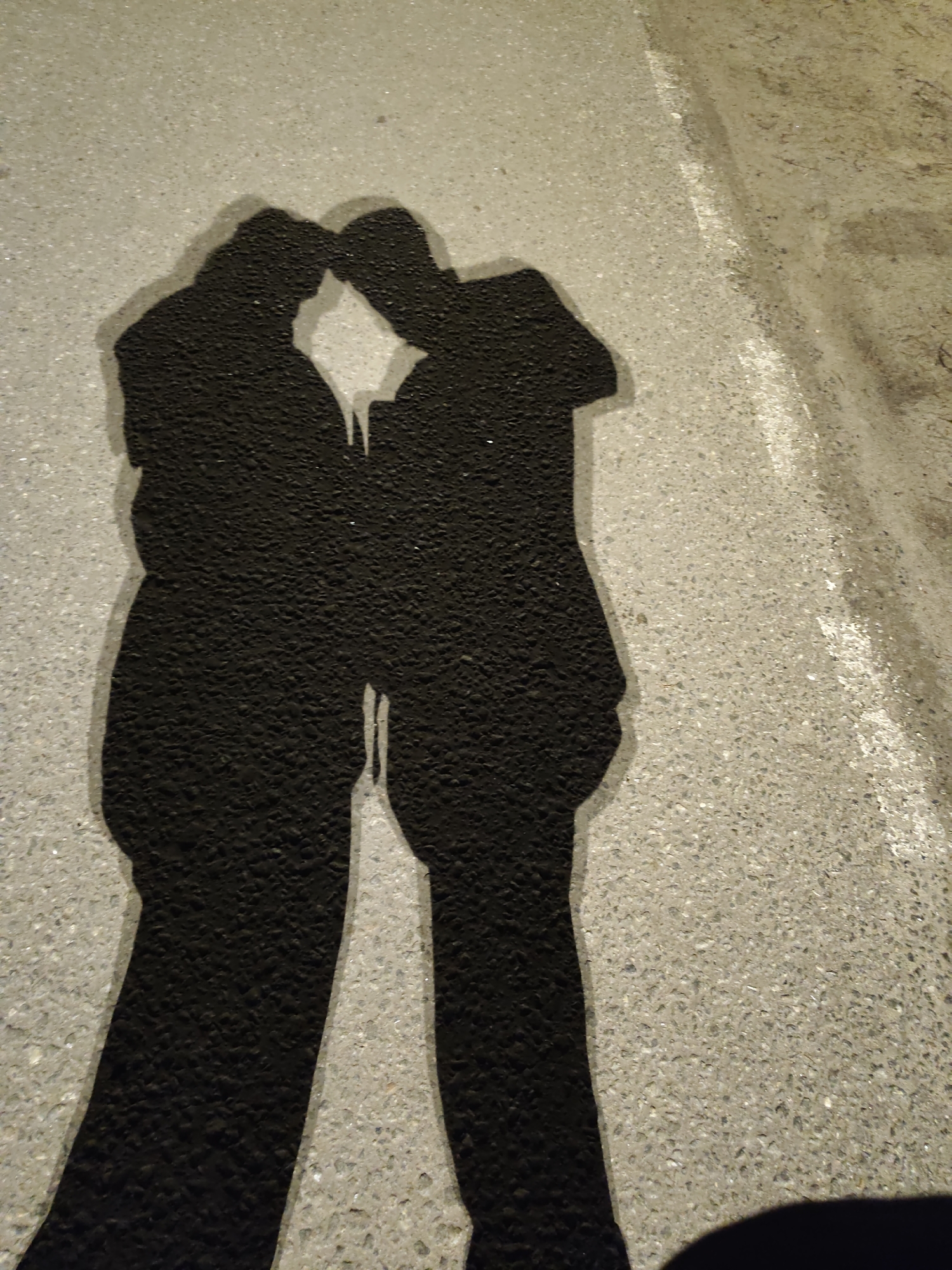 The image depicts the shadows of two individuals on a paved surface, likely a sidewalk or road, marked with a white line. The shadows suggest that the two people are standing close to each other, possibly embracing or with their heads touching. The shadows obscure any details about their individual appearances. The surface is light grey with a coarse texture. The setting appears to be outdoors and illuminated by an unseen light source that is casting the shadows. The presence of the white line suggests that the location may be near a street. The city of Kratovo, Russia is not evident in the image.