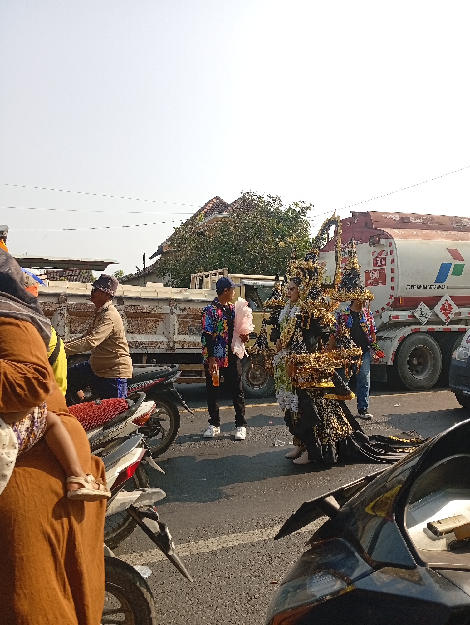 In Sarang, Indonesia, a street scene unfolds with a large Pertamina tanker truck, identified by its logo and red and white coloring, dominating the background. The truck, with license plate B 9365 UFU, displays hazard symbols and a speed limit of 60 km/h. To the left of the truck, a yellow truck with a white cargo bed is visible, alongside a man in a brown shirt and bucket hat riding a motorcycle.
In the foreground, two individuals in elaborate black and gold traditional costumes stand on the asphalt road, holding what appear to be decorative items. One person in the costume is holding a pink object. To their left, a person in a colorful, patterned shirt holds a drink. A woman in a brown headscarf and dress carries a child on her back, the child wearing sandals and a patterned skirt. Several motorcycles are parked or in motion on the road, including one with a red seat. The environment suggests a sunny day with clear skies and houses with tiled roofs visible in the distance. The overall scene captures a moment of local activity, possibly a parade or cultural event, on a public street.