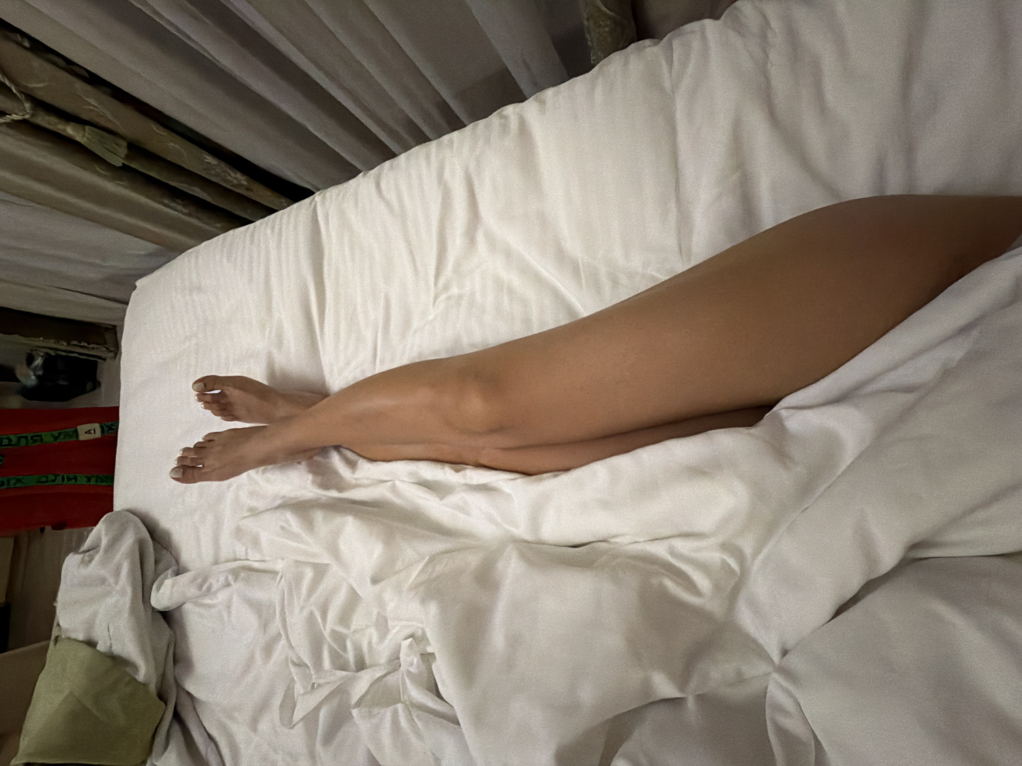 A person's legs are visible, covered by white bedding on a bed. The person's feet, with white-painted toenails, are crossed at the ankles. The environment appears to be a bedroom, with curtains visible in the background. The location in Zelenogradsk, Russia cannot be determined from the image.