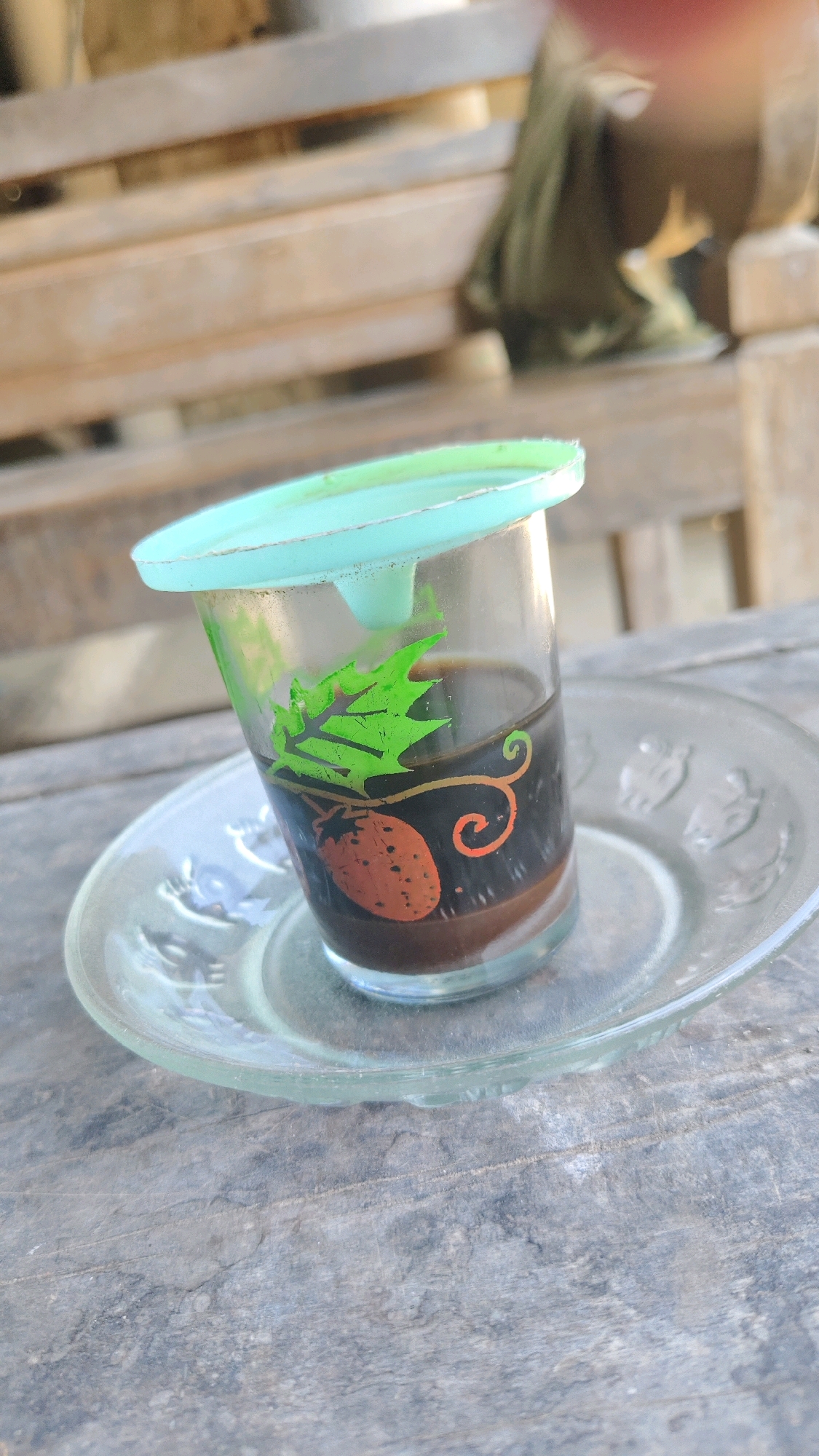 A clear glass cup filled with dark liquid, likely coffee, sits on a clear glass saucer. The cup has a decorative pattern featuring a strawberry, green leaves, and an orange swirl. A translucent light blue lid is placed on top of the cup. The cup and saucer are positioned on a weathered wooden surface, possibly a table or bench. In the blurred background, wooden slats suggest a seating area. The overall scene appears to be outdoors, possibly in a rustic or casual setting in the city of Galis, Indonesia.