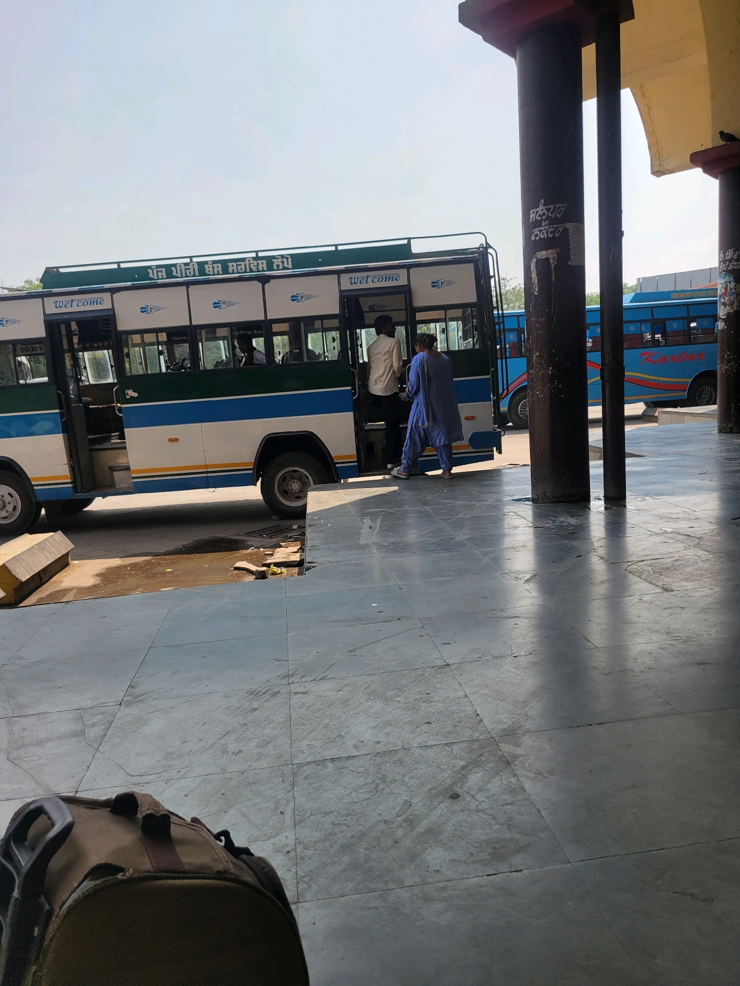 The image depicts a bus stop in Jagraon, India, under a clear sky. A bus with green and blue stripes is parked at the station, with passengers boarding. A woman in a blue salwar kameez is entering the bus, followed by a man in a light-colored shirt. Another bus, blue in color with "Kartar" written on its side, is visible in the background. The bus stop has a covered waiting area supported by large pillars, some of which have markings in Punjabi. The floor is tiled with grey stone. In the foreground, a duffel bag rests on the tiled floor. The scene captures a moment of transit at a busy bus station.