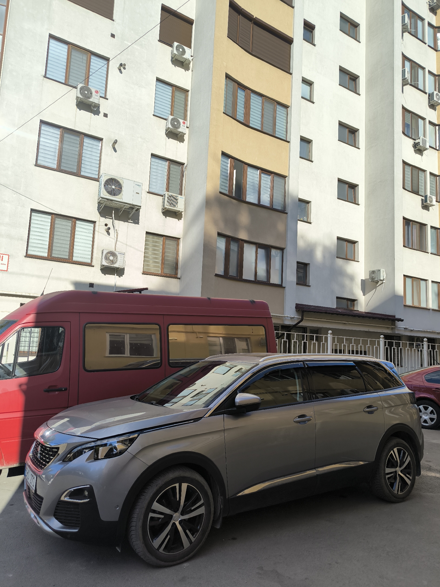 The image captures a sunlit urban street scene in Chișinău, Moldova. A silver Peugeot 3008 SUV is parked in the foreground, facing left, with its front end angled towards the viewer. To the left of the SUV is the side of a red van. Behind the vehicles, a modern apartment building stands, its facade a mix of white and pale yellow. Multiple windows with wooden frames and roller blinds are visible on the building's exterior, along with several air conditioning units. The street surface appears to be asphalt. The lighting suggests it is daytime, with shadows cast by the building onto the vehicles and the ground.