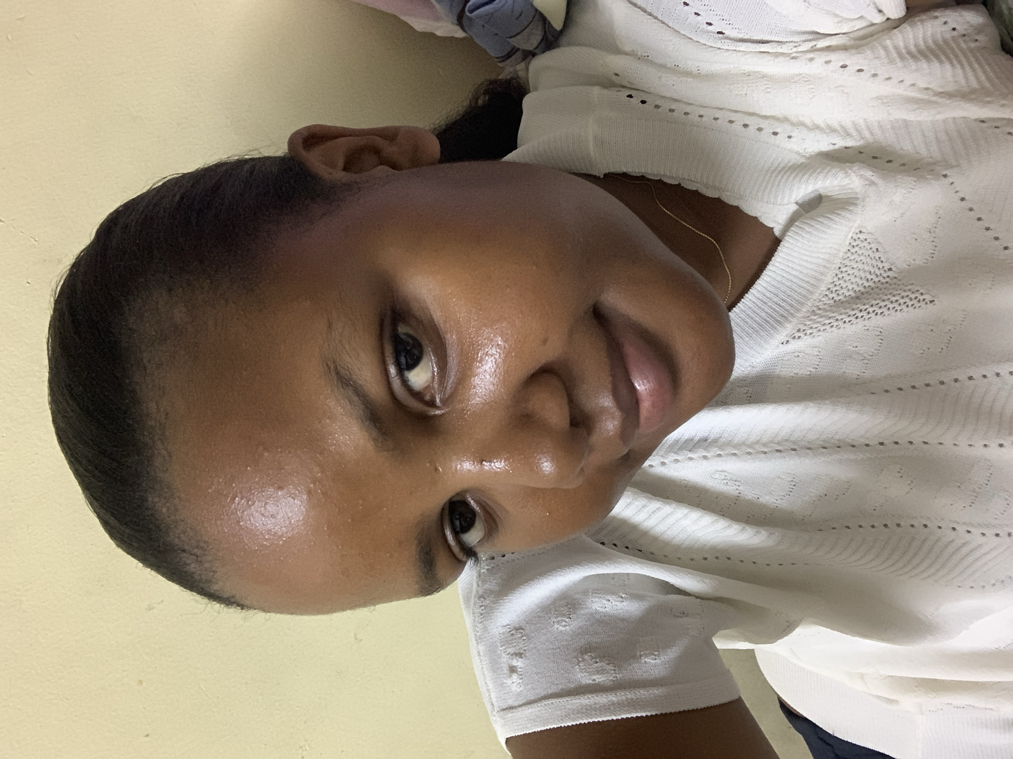A close-up shot focuses on a dark-skinned woman with short, dark hair, wearing a white, knitted, round-necked top. She is tilted at an angle, looking directly at the camera with a slight smile. A thin gold necklace is visible at her neck. The background is a plain, pale yellow wall. The location is not discernible from the image.