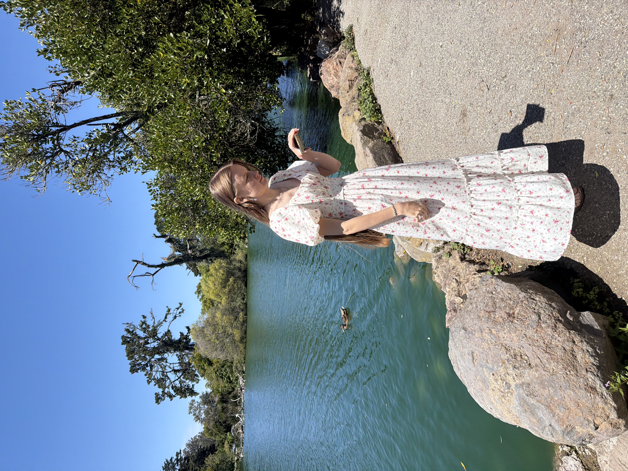 A young woman stands on a gravel path next to a lake in San Francisco, California. She is wearing a white floral print dress with puffy sleeves and a tiered skirt. The dress is white with small pink flowers. She has long, light brown hair and is looking down at her hand, which is holding a small object, possibly a phone. In the background, a tranquil lake with green water stretches out, with a duck swimming on its surface. Lush green trees and a bright blue sky form the backdrop. The path is bordered by large rocks and vegetation. The overall scene is bright and sunny, suggesting a pleasant day outdoors.