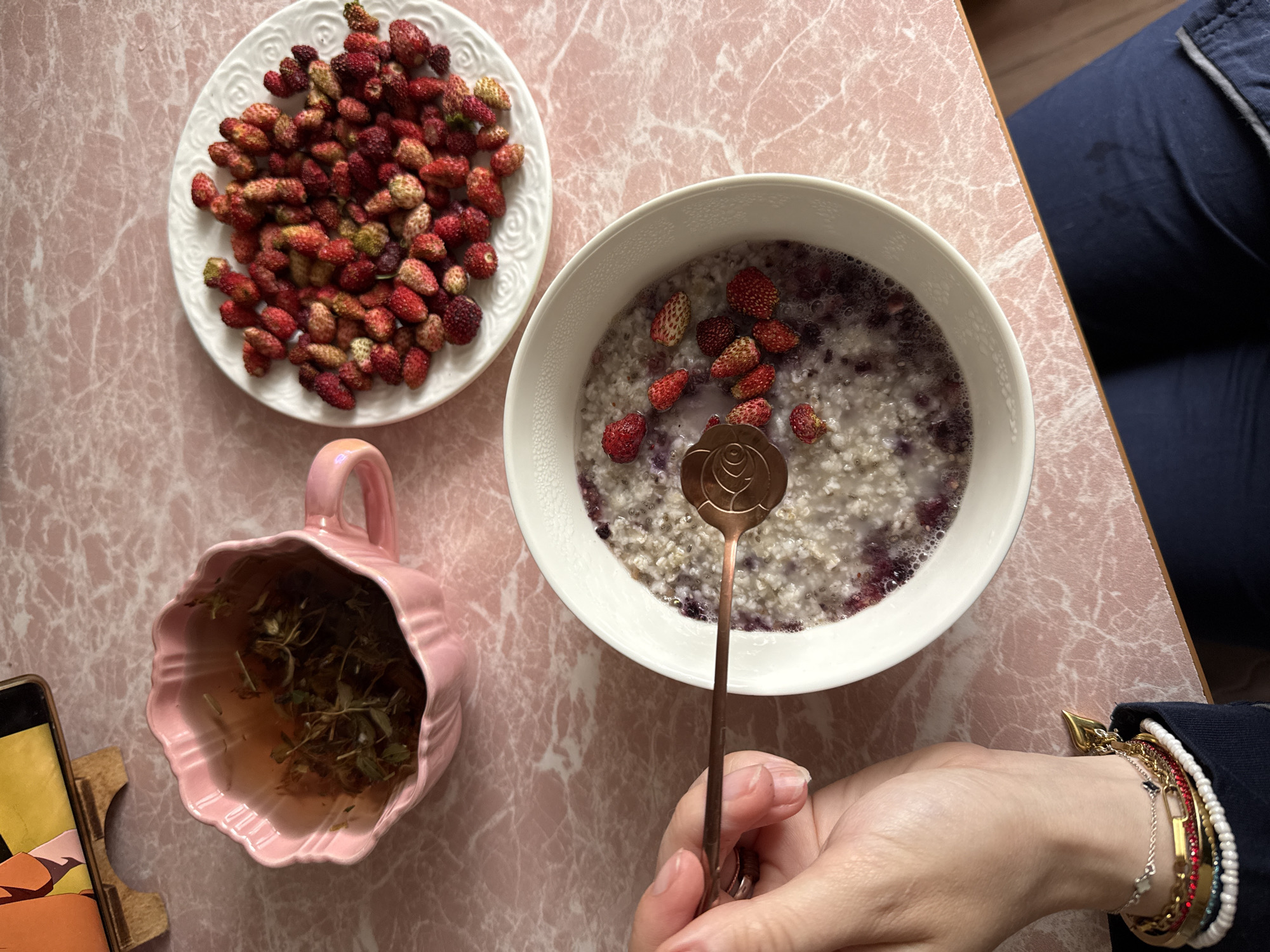 The image shows a top-down view of a breakfast setting on a pink marble-patterned surface. In the foreground, a white bowl contains oatmeal topped with blueberries and small wild strawberries. A copper spoon with a rose design is partially submerged in the oatmeal. To the left of the bowl, a hand with several bracelets is visible, holding the spoon. To the upper left of the bowl, a pink, scalloped teacup is filled with herbal tea. To the right of the teacup and above the bowl, a white plate is piled high with wild strawberries. A phone on a wooden stand is visible in the upper left corner, displaying an orange and yellow graphic. The overall scene suggests a light and healthy breakfast.