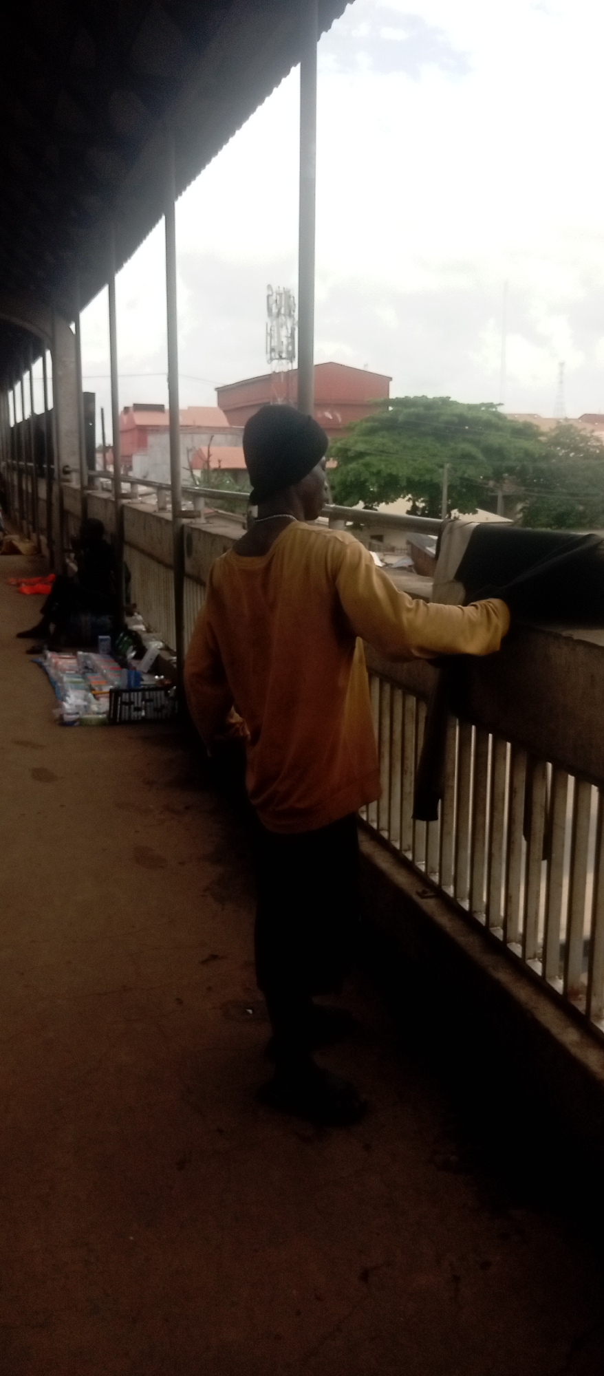 The image depicts a person standing on an elevated walkway, likely a pedestrian bridge, in Idemili, Nigeria. The person is wearing dark pants, a light orange long-sleeved shirt, and a dark head covering. They are leaning on the railing of the bridge, looking out at the cityscape. Other people are present nearby. Merchandise is set up next to the walkway, indicating possible informal trading. The environment includes buildings, trees, and communication towers in the background, under an overcast sky.