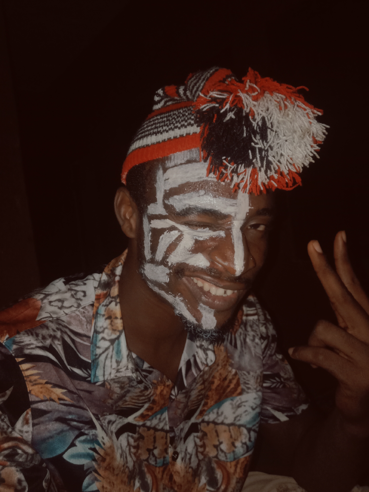 A young man from Onitsha, Nigeria, is featured in a close-up shot, adorned with white face paint in striking patterns across his forehead, cheeks, nose, and chin, suggesting involvement in a cultural event or celebration. He wears a vibrant, knitted cap with red, black, and white patterns, topped with decorative tassels of similar colors. His attire includes a short-sleeved shirt with a multi-colored abstract or floral design. The man is smiling broadly and winking with his left eye, conveying joy and playfulness, while his right hand forms a V-sign. The background is dark, indicating the photo was likely taken at night or in a dimly lit indoor setting. No text is visible in the image.