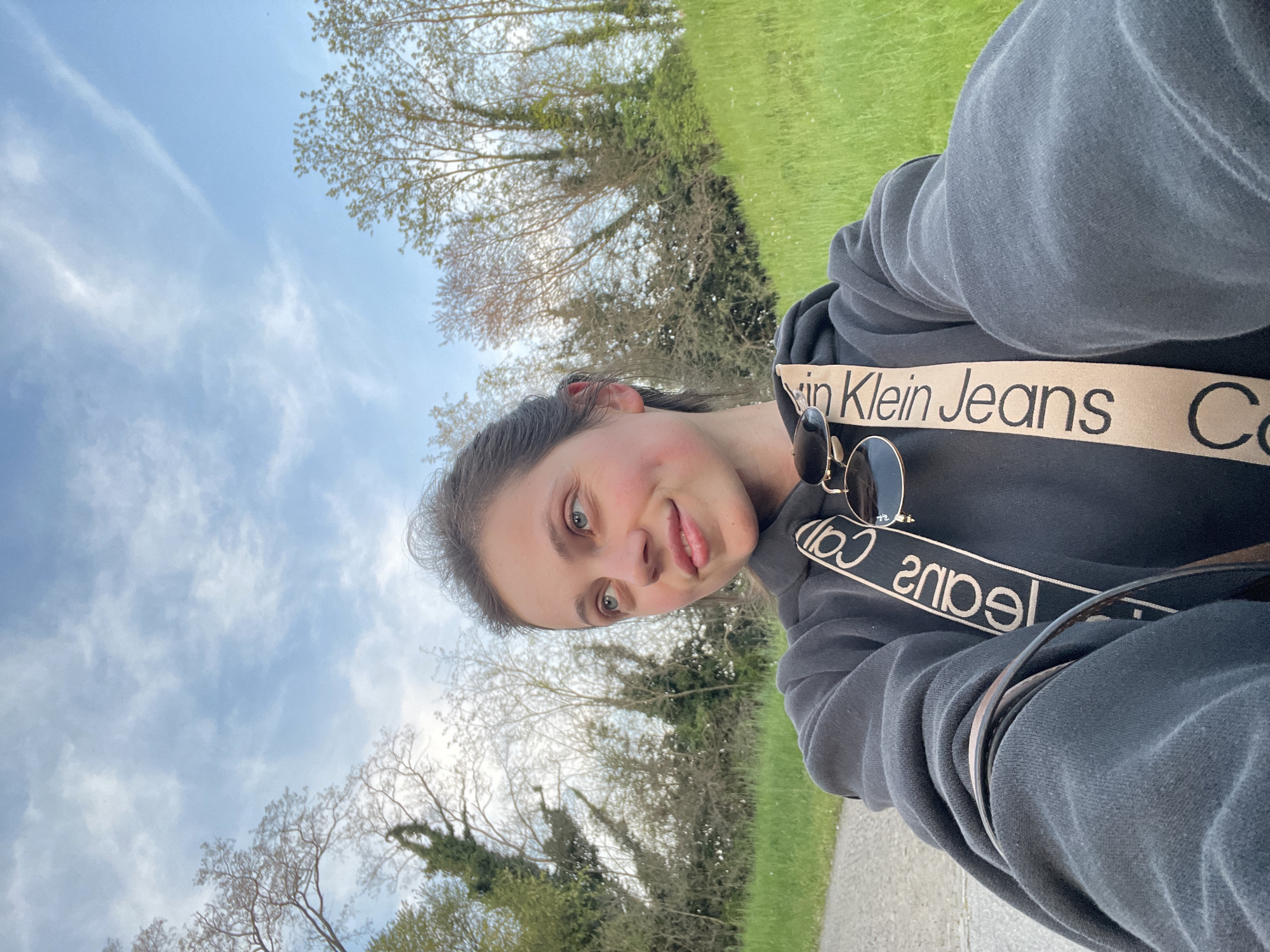 A selfie of a young woman is taken outdoors. The background reveals trees, a field, and a blue sky with sparse clouds. The woman wears a grey hoodie with a Calvin Klein Jeans band. Sunglasses are positioned on her hoodie. The photo's angle gives a slightly tilted perspective of the surrounding environment. The image might be from a place near Case / Scjasis, Italy, as per location query, but lacks any defining landmarks or geographical features from the area.
