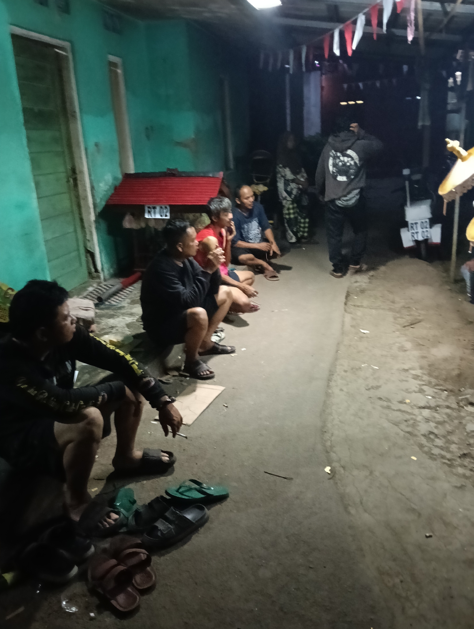 The image depicts an outdoor gathering of men in what appears to be a residential area in Tasikmalaya, Indonesia. The scene is dimly lit, suggesting it is evening or night. Several men are seated on the ground in a row along a wall painted teal. Some are looking towards the right side of the frame, while others are looking down. One man in the foreground on the left is smoking a cigarette and holding a phone. Several pairs of sandals are scattered on the ground near the seated individuals. In the background, a person wearing a grey hooded sweatshirt with a graphic on the back is standing and appears to be interacting with others out of frame to the right. A motorcycle is also visible in the background, with license plates displaying "RT 02". Decorative flags are strung overhead. The environment appears to be a walkway or alleyway with a rough concrete surface.