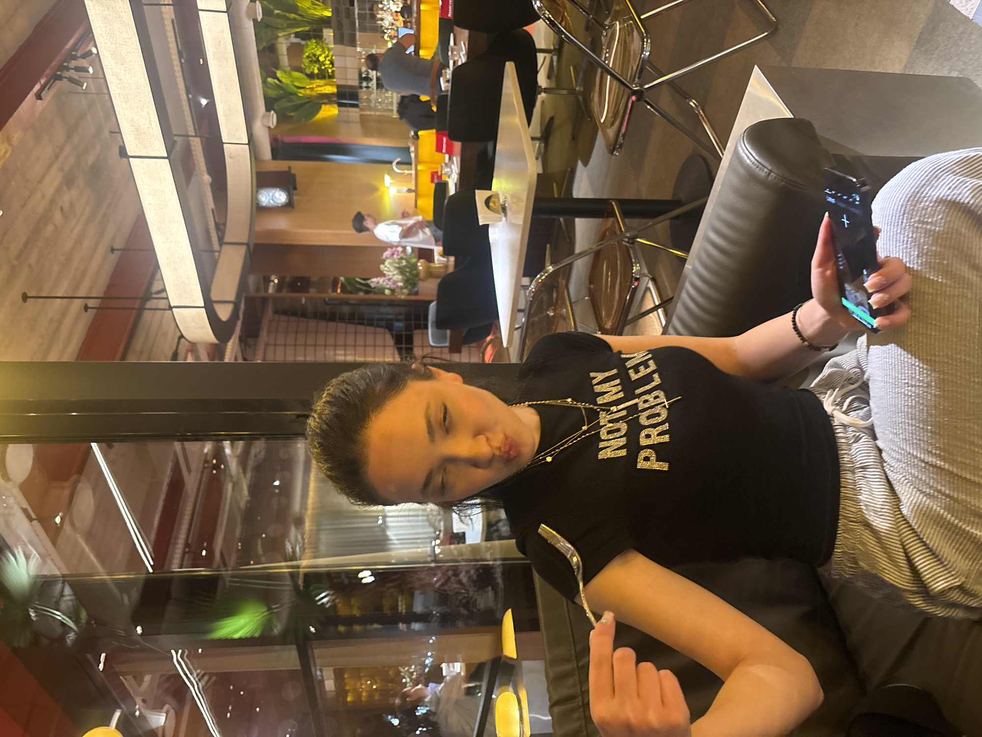 The image captures a playful moment inside a modern restaurant or cafe, likely in the evening given the artificial lighting. The main subject is a woman with dark hair, seated on a black leather couch or booth. She is looking towards the camera with a pouting, kissing face. She wears a black t-shirt emblazoned with the sparkly gold text "NOT MY PROBLEM" and a gold necklace. In her right hand, she holds a smartphone, its screen illuminated, and in her left, she holds a fork. Her lower body is partially visible, dressed in light-colored, striped pants.
The background reveals a bustling interior with warm, ambient lighting. Various tables and chairs, some with clear or metallic frames, are visible. There's an elongated, oval-shaped light fixture overhead, providing illumination. Further back, a bar area is visible with bottles, glasses, and what appears to be staff or other patrons. One person in the background, possibly a waiter, is wearing a white shirt and moving around. Plants are strategically placed throughout the space, adding greenery. Reflections on glass or polished surfaces suggest a window or mirror, mirroring parts of the interior. The overall setting is chic and contemporary. The location context provided is Yerevan, Armenia.