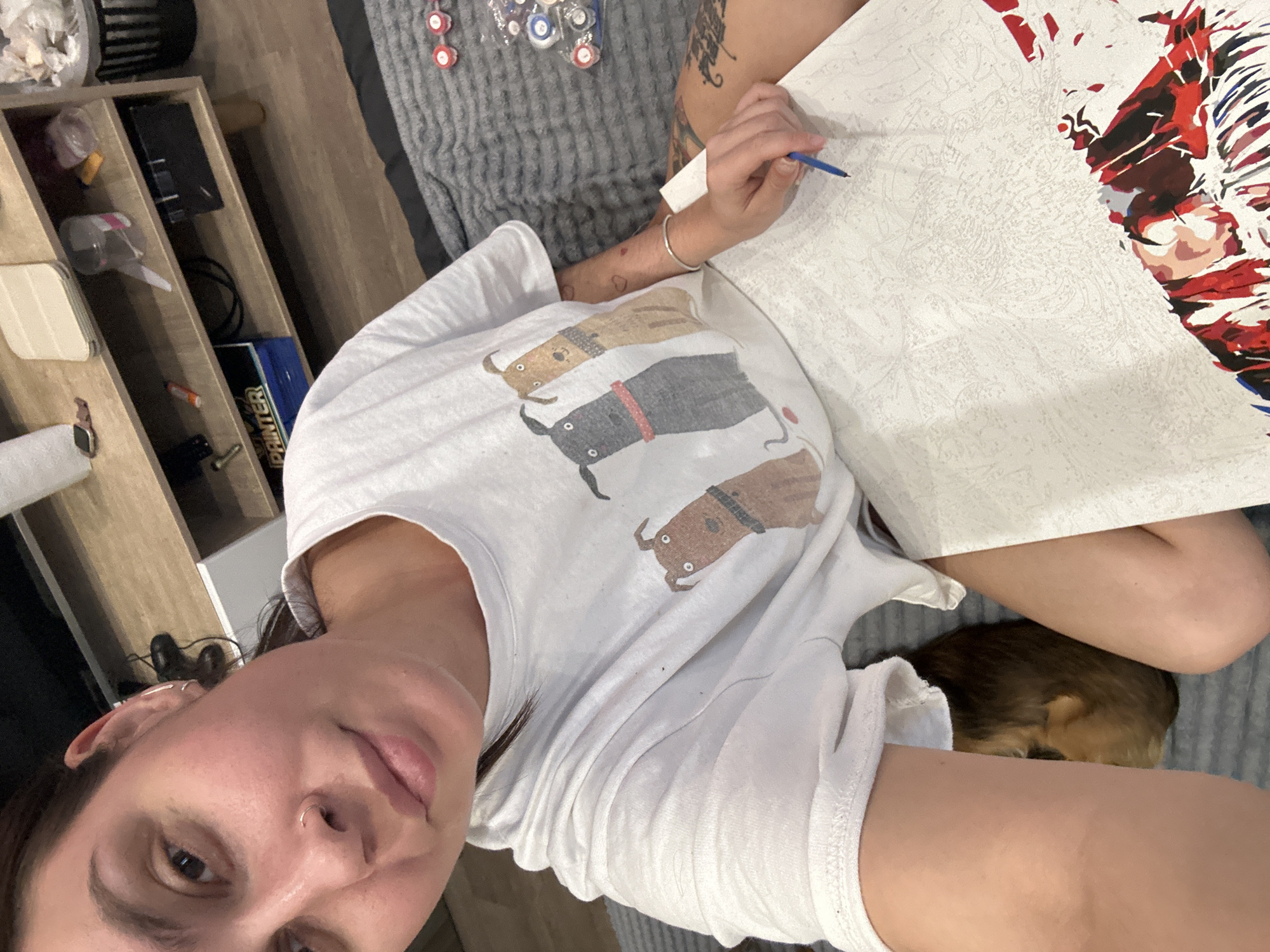 A woman is seated on a gray, textured surface, likely a bed or couch. She is wearing a white t-shirt with a cartoon graphic of three dogs. She is holding a blue paint brush and appears to be working on a paint-by-numbers canvas that features an image with red, white, and blue colors. A small, brown and black dog is curled up near her legs. A wooden shelving unit is visible in the background, with various items on the shelves. The location in Izhevsk, Russia cannot be determined from this image.
