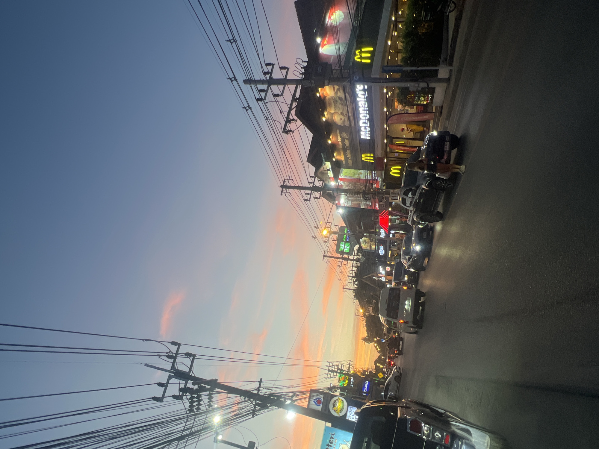 The image depicts a street scene in the city of Ko Samui District, Thailand, during sunset. The sky displays a gradient of blue to pink and orange hues, with wispy clouds. A street is lined with buildings, including a McDonald's restaurant illuminated with its signature golden arches and signage. Power lines are prominently visible, crisscrossing the sky above the street. Several vehicles, including cars and vans, are on the road, suggesting moderate traffic. A woman is seen standing on the sidewalk near a black pickup truck. The overall atmosphere is that of a bustling urban environment at dusk.