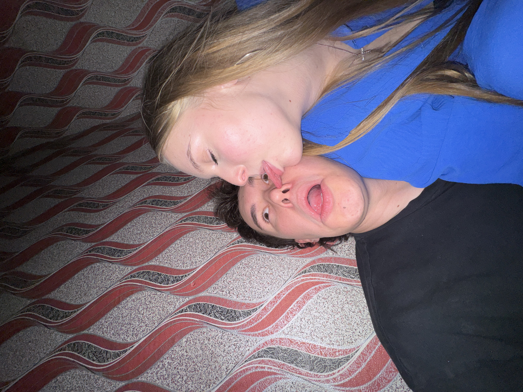 A young woman with long blonde hair, wearing a bright blue jacket, is leaning in to kiss a young man on the cheek. The man, with dark hair, is looking upwards with his mouth open and his tongue sticking out. They are positioned in front of a wall with a bold, wavy red and grey patterned wallpaper. The scene appears to be indoors, possibly in a home or establishment with distinctive decor, and is likely located in the city of Perm, Russia.