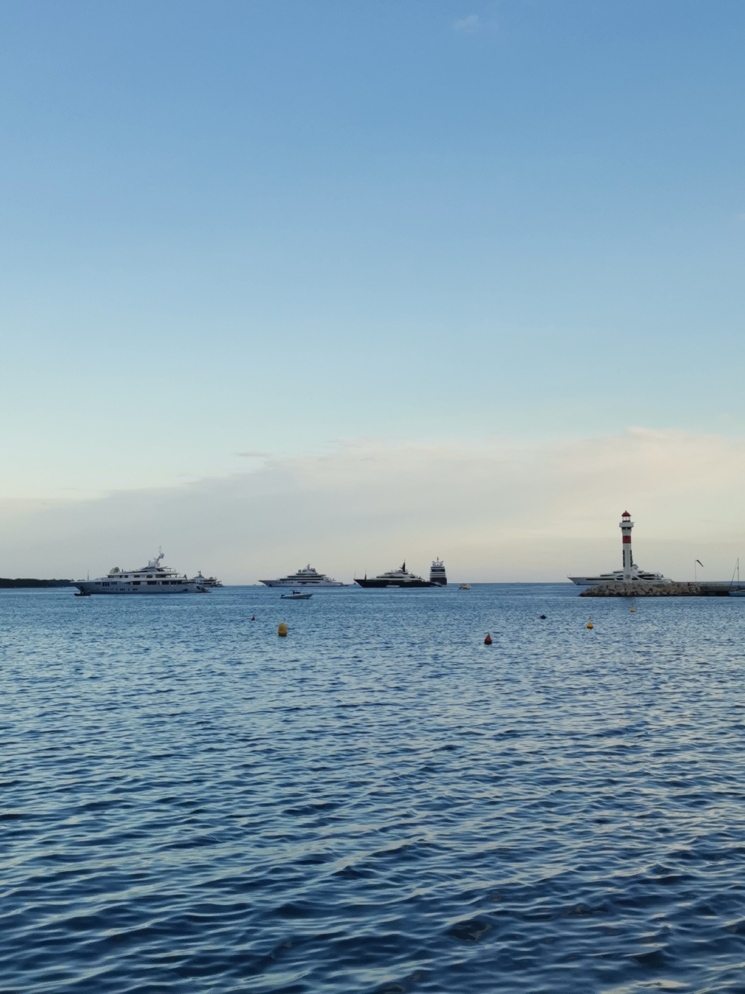 The image shows a seascape under a blue sky, possibly at dusk, featuring several yachts and a lighthouse. Three large yachts are visible in the middle ground, positioned at varying distances from the viewer. The lighthouse stands on a rocky breakwater to the right. Small yellow and red buoys float on the surface of the water. The location is likely near a coastline with maritime activity, in an unknown city. The scene conveys a sense of tranquility and maritime activity.