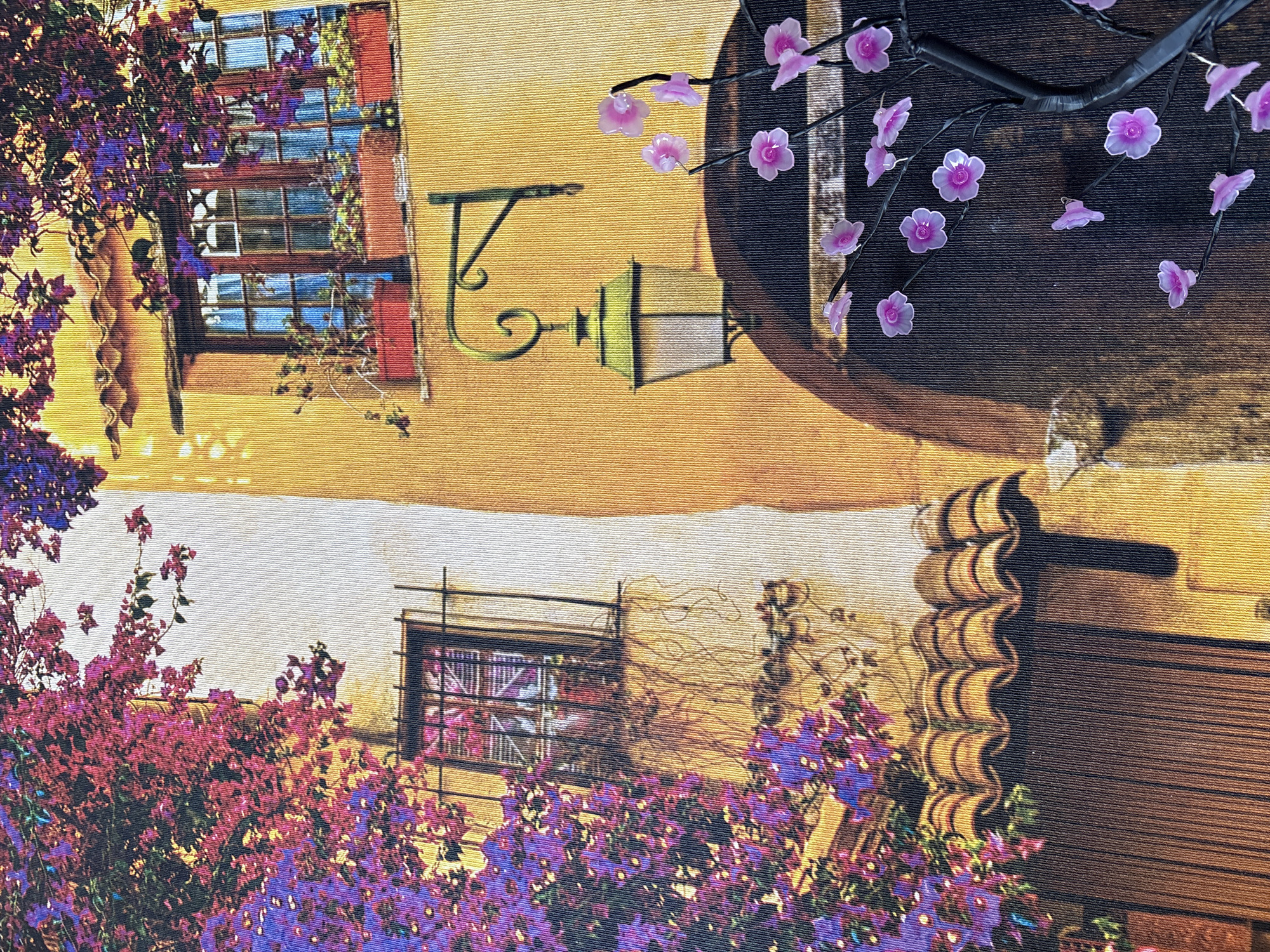 This is a photograph of a building with a stucco exterior painted in warm, sun-kissed tones. Vibrant pink and purple bougainvillea flowers cascade down the left side of the building, creating a lush canopy over a window with a barred gate. The window is framed by dark wood and has a row of red planters overflowing with greenery. On the right side of the building, a second window is visible, also adorned with red planters. Below this window, a vintage-style street lamp with a green lantern hangs from an ornate black metal bracket. In the foreground, a decorative tree with delicate pink flowers is partially visible. The image appears to be a close-up, possibly of a painting or a textured fabric print, rather than a direct photograph of a street scene. The location of Georgiyevsk, Russia, is not discernible from the visual elements of the image.