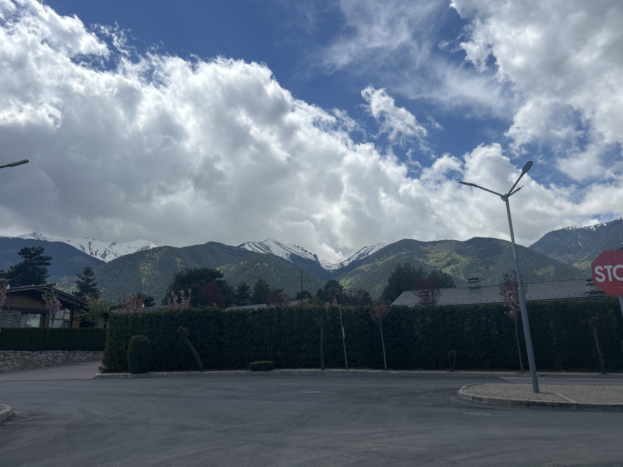 The image depicts a street scene in an unknown location in Bulgaria. The sky is predominantly blue with scattered white clouds. In the background are green mountains with patches of snow on their peaks. Closer to the viewer are buildings and lush green hedges bordering the street. A street light is visible on the right side of the road, along with a red stop sign partially obscured by the pole. The street is paved with dark asphalt, and the general atmosphere suggests a quiet, possibly residential, neighborhood.