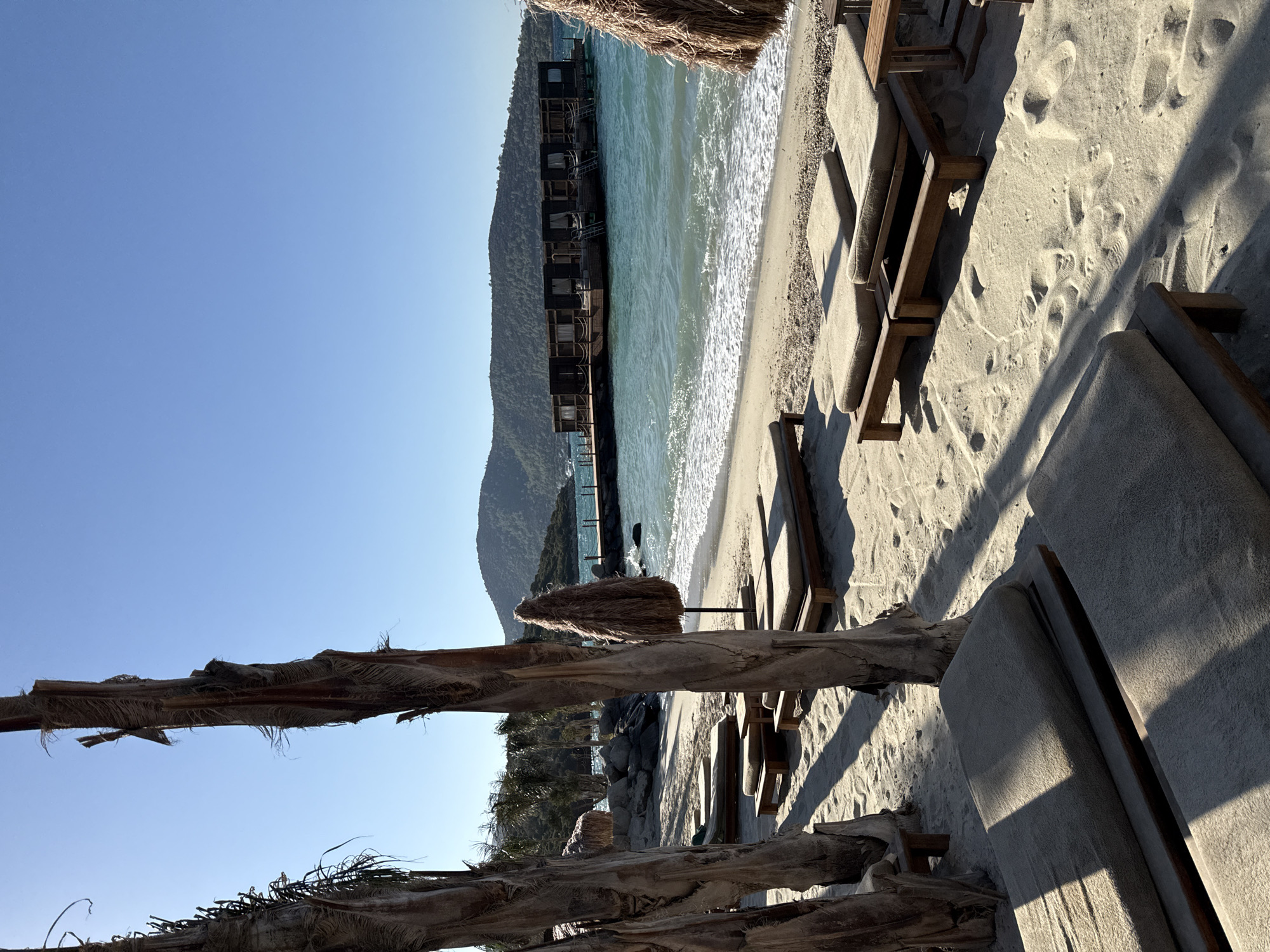 A serene beach scene in Bodrum, Turkey, bathed in the warm glow of sunlight. In the foreground, rows of empty, cushioned lounge chairs are neatly arranged on the soft, sandy beach, casting long shadows from the nearby palm trees. The palm trees themselves, with their weathered, fibrous trunks, stand tall, framing the view.
Further back, a beach umbrella with a straw canopy adds a touch of tropical ambiance. Beyond the immediate beach area, the turquoise waters of the Aegean Sea gently lap against the shore, creating white foam. In the middle ground, a wooden pier extends into the water, leading to a unique structure of interconnected, dark wooden cabins or bungalows built on pontoons. These floating accommodations are a distinctive feature of the scene.
In the background, rolling hills covered in lush greenery rise against a clear, bright blue sky. The overall impression is one of tranquility and relaxation, typical of a luxury seaside resort in Bodrum. There are no people visible in the image. The sand is marked with footprints, suggesting recent activity. The weather is clear and sunny, with no clouds.