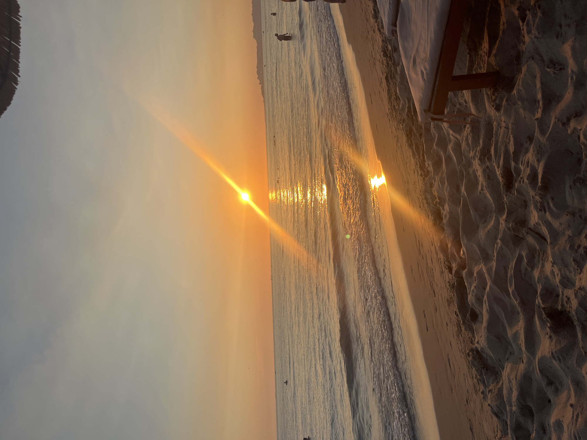 The image depicts a tranquil sunset scene at a beach, likely located in the city of Bashkia Durrës, Albania. The sun, a brilliant orange orb, is setting over the calm, shimmering sea, casting a warm glow across the sky and water. Sunbeams dramatically streak across the frame, reflecting brightly on the ocean's surface. Gentle waves lap at the sandy shore, which is adorned with the textured patterns of sand. In the foreground, two empty lounge chairs with white cushions are positioned on the sand, awaiting visitors. Several people are visible in the water, some standing and others seemingly engaged in swimming or wading, indicating a leisurely evening atmosphere. In the distance, a hazy coastline with some structures can be discerned. The overall impression is one of serenity and natural beauty, characteristic of a coastal sunset.
