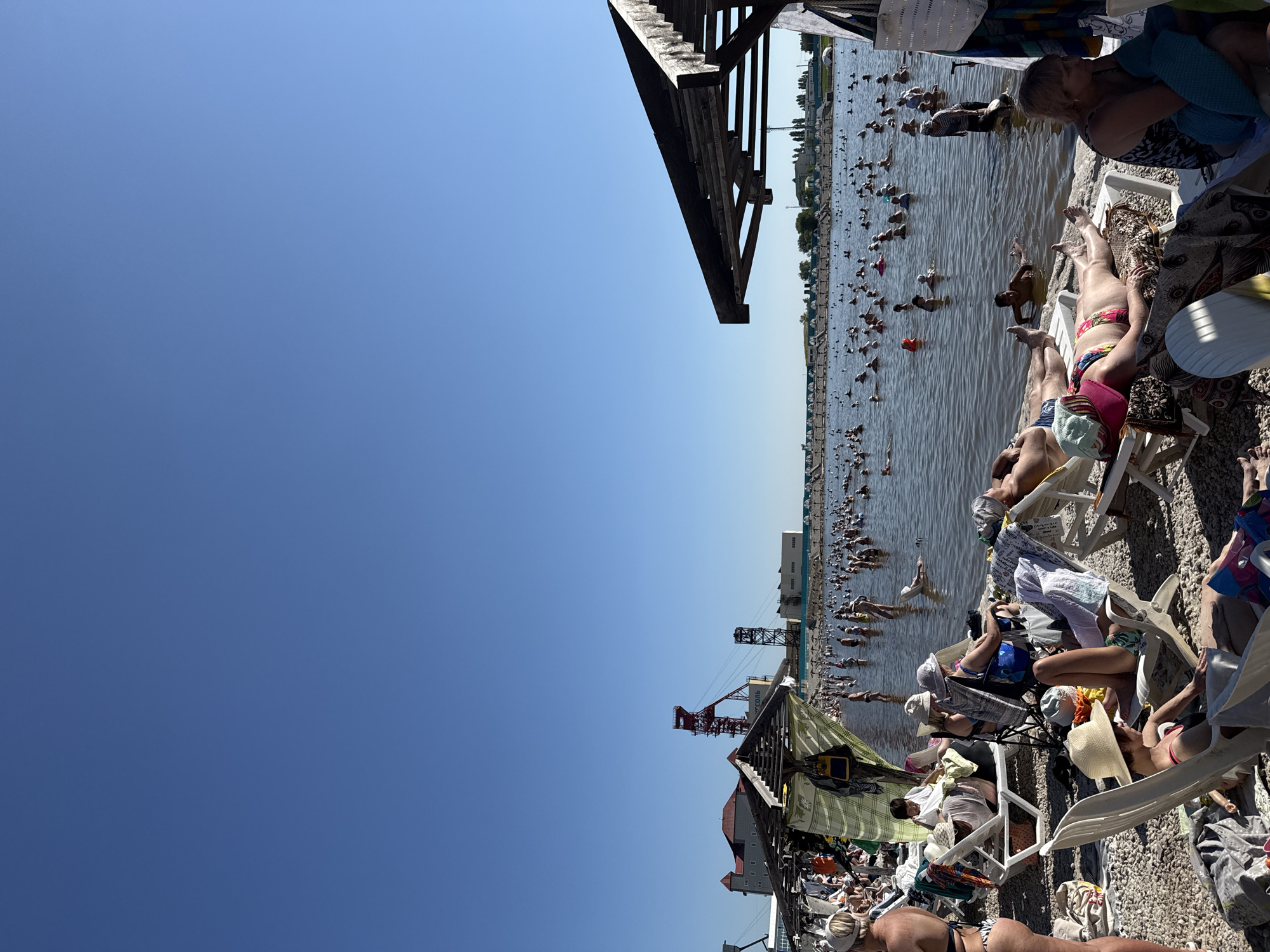 This is a daytime, eye-level, wide shot of a crowded beach in Sol-Iletsk, Russia, likely on a saline lake. The sky is clear and blue. In the foreground, numerous beach chairs are occupied by sunbathers, some of whom are wearing hats and swimwear. The beach is covered with pebbles. To the left of the frame, part of a covered structure with hanging clothes is visible. In the middle ground and background, a vast number of people are wading and swimming in the shallow, murky water. In the distance, industrial structures, including a tall red tower with a yellow accent, are visible on the shore, along with a long building and a row of small umbrella-like structures. The overall scene depicts a popular recreational spot with many visitors enjoying the water and sun.