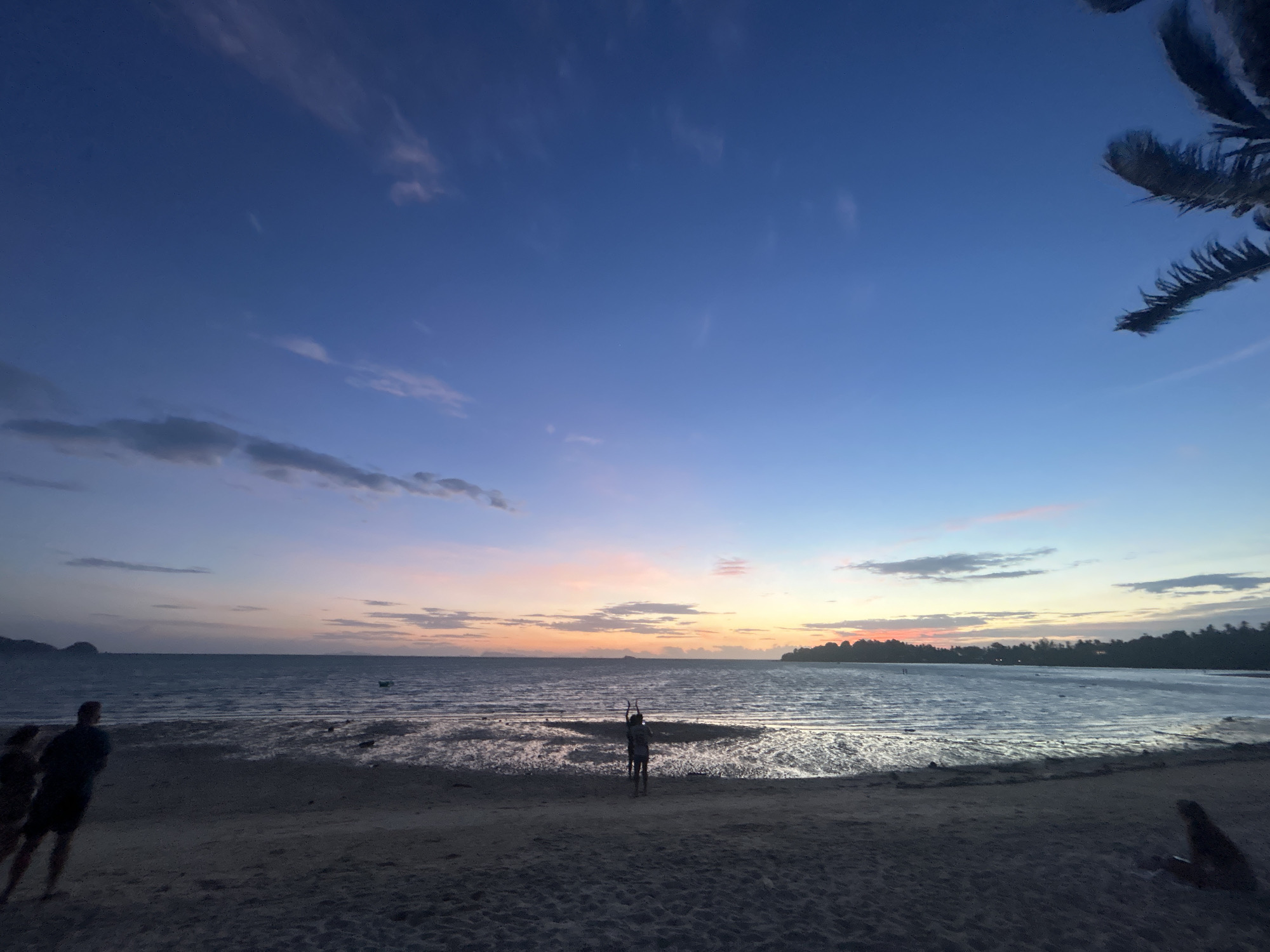 The image depicts a beach scene at dusk in the Ko Pha-ngan District, Thailand. The sky is a gradient of deep blue to lighter blue with streaks of pink and orange clouds near the horizon, indicating sunset. The sea is calm with gentle waves reflecting the sky. The foreground is a sandy beach with footprints visible. In the mid-ground, a person stands facing the water with their arms raised, seemingly in a pose of contemplation or exercise. To the left, two figures, partially obscured by shadow, are walking on the beach. A palm tree frond is visible in the upper right corner, silhouetted against the sky. The overall atmosphere is serene and tranquil.