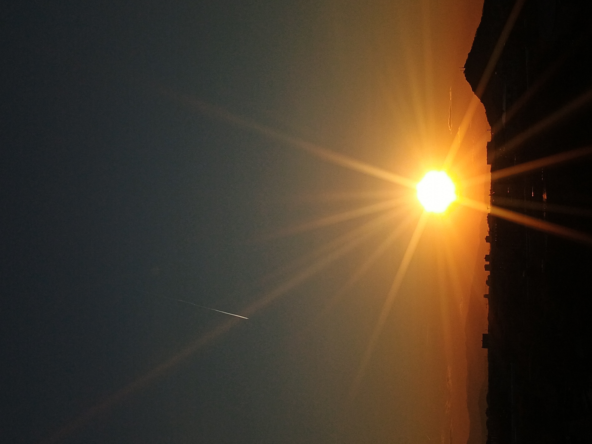 The image captures a sunset in Plovdiv, Bulgaria. The sun, a bright orb with radiating beams, is positioned near the horizon, casting a warm orange glow. Silhouetted against this light is the city skyline with buildings, suggesting a location within or near an urban area. A contrail from an airplane is visible in the dark sky, adding to the scene's sense of distance.