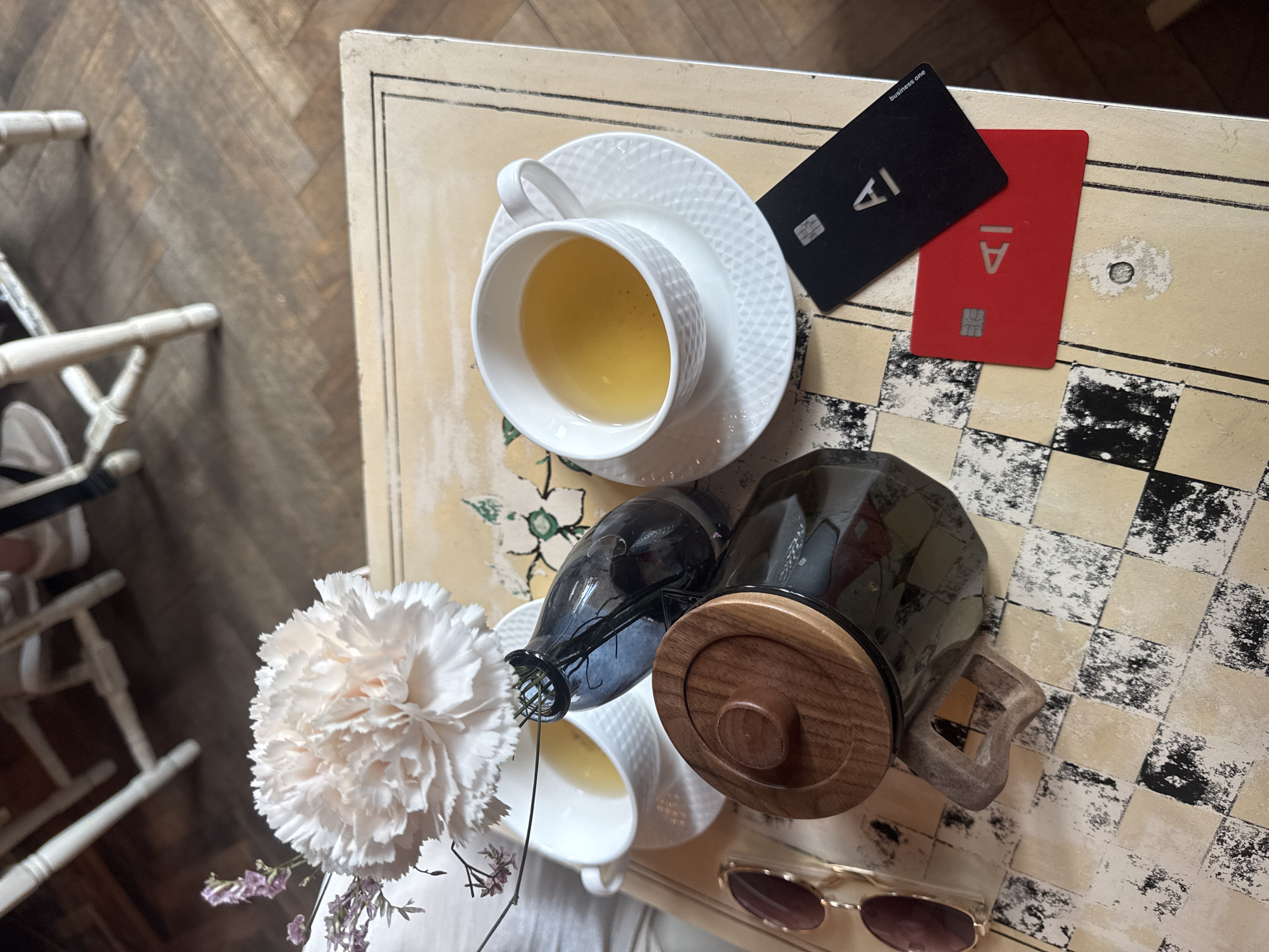 This is a high-angle, medium shot of a table setting at a cafe in Saint Petersburg, Russia, on a warm, overcast day. The table, with a distressed cream-colored surface featuring a black and beige checkered pattern, appears to be outdoors, possibly in a courtyard, with wooden floorboards visible in the background.
The foreground is occupied by several objects. A large, light-pink carnation with delicate purple sprigs is positioned towards the upper left. To its right, a white teacup with a textured pattern sits on a matching saucer, filled with a golden-yellow liquid, likely tea. Next to the teacup is a dark, translucent glass bottle, possibly a decanter or vase, containing thin green stems. In front of this, a glass teapot with a wooden lid and handle is partially visible. To the left of the teapot, a pair of sunglasses with brown lenses and gold frames rests on the table.
Dominating the lower right portion of the image are two credit cards: one red and one black. Both cards feature a white "AI" logo. The black card also has the text "business one" in white on its lower right corner. The table surface is uneven and appears to have some wear and tear, with a circular area of chipped paint near the bottom center.
There are no people clearly visible in the frame, though blurred shapes in the background suggest seating and possibly patrons of the cafe. The lighting is soft and diffused, indicating natural light. The overall impression is one of a relaxed, sophisticated cafe experience.