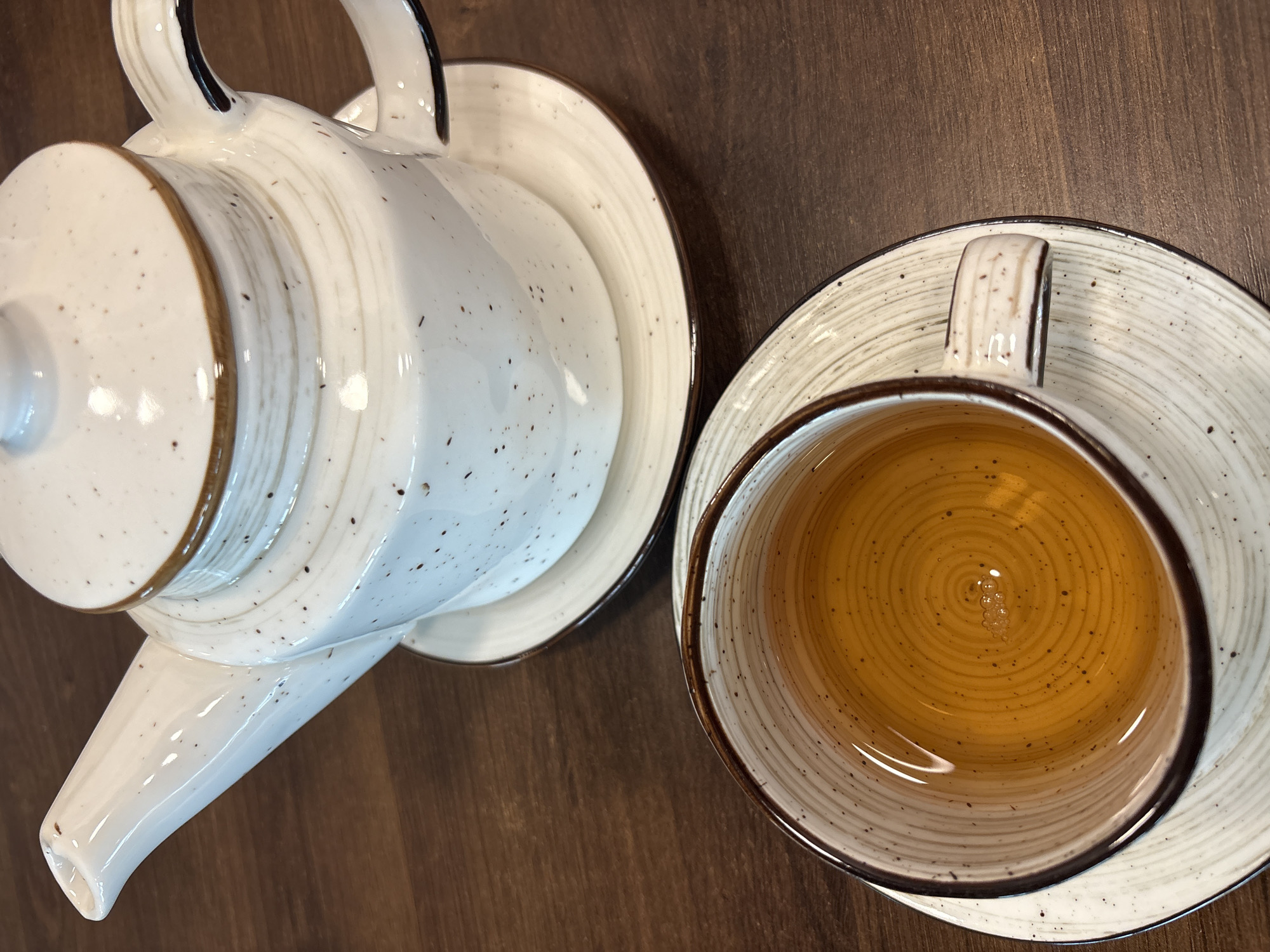 An overhead view captures a ceramic teapot and a teacup on a dark brown wooden table in the city of Chaikovsky, Russia. The white ceramic teapot features irregular brown speckles and a brown line along the rim of its lid and handle, resting on a matching saucer with a brown rim. Below it, a white ceramic teacup, similarly patterned with brown speckles and a brown rim, holds an amber-colored liquid with small bubbles visible on its surface. The teacup is placed on a matching saucer. The wooden table exhibits a visible grain pattern. No individuals are present in the scene.