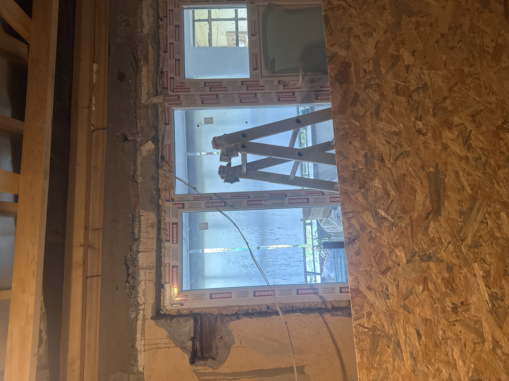 This image captures an interior construction scene in Sarnevo, Bulgaria, depicting a newly installed window and unfinished structural elements.
The upper portion of the frame reveals exposed wooden ceiling joists running horizontally, with a dark void visible between them, indicating an unfinished ceiling or floor structure above. Below the joists, a roughly textured concrete or masonry wall is present. This wall shows evidence of demolition or ongoing work, with irregular edges, exposed aggregate, and patched areas. A section of rebar or a dark pipe is visible within a cavity on the left side of the wall, immediately adjacent to the window frame. Expanding foam insulation is visible along the top perimeter of the window and in gaps within the upper wall. A single black cable descends from the upper wooden structure into the wall.
The central feature is a white multi-pane PVC window unit. The window frame displays repetitive "HAMMER" branding in red and blue text on a white background. The window unit is sealed into a rough opening with visible expanding foam insulation around its perimeter. Through the window, looking outwards, an exterior environment is discernible. The left pane shows a slightly obscured view, possibly of foliage or an outdoor scene, with what appears to be a light-colored blind or curtain on the interior side of the glass. The central panes frame a silver-colored A-frame ladder, positioned upright, its top visible through the glass. The right pane offers a view of another structure, possibly part of an adjacent building or another window, with a discernible grid pattern. Below this, a section of a greenish-grey surface is visible. Two parallel white electrical wires traverse horizontally across the lower central panes of the window.
The lower portion of the image is dominated by a sheet of Oriented Strand Board (OSB), which acts as a barrier or surface covering, obscuring the floor or lower wall sections beneath the window. The OSB shows the characteristic irregular wood chip pattern.
No individuals are present in the scene. The lighting suggests a combination of interior artificial light and natural light filtering through the window, indicating an active construction or renovation environment.
