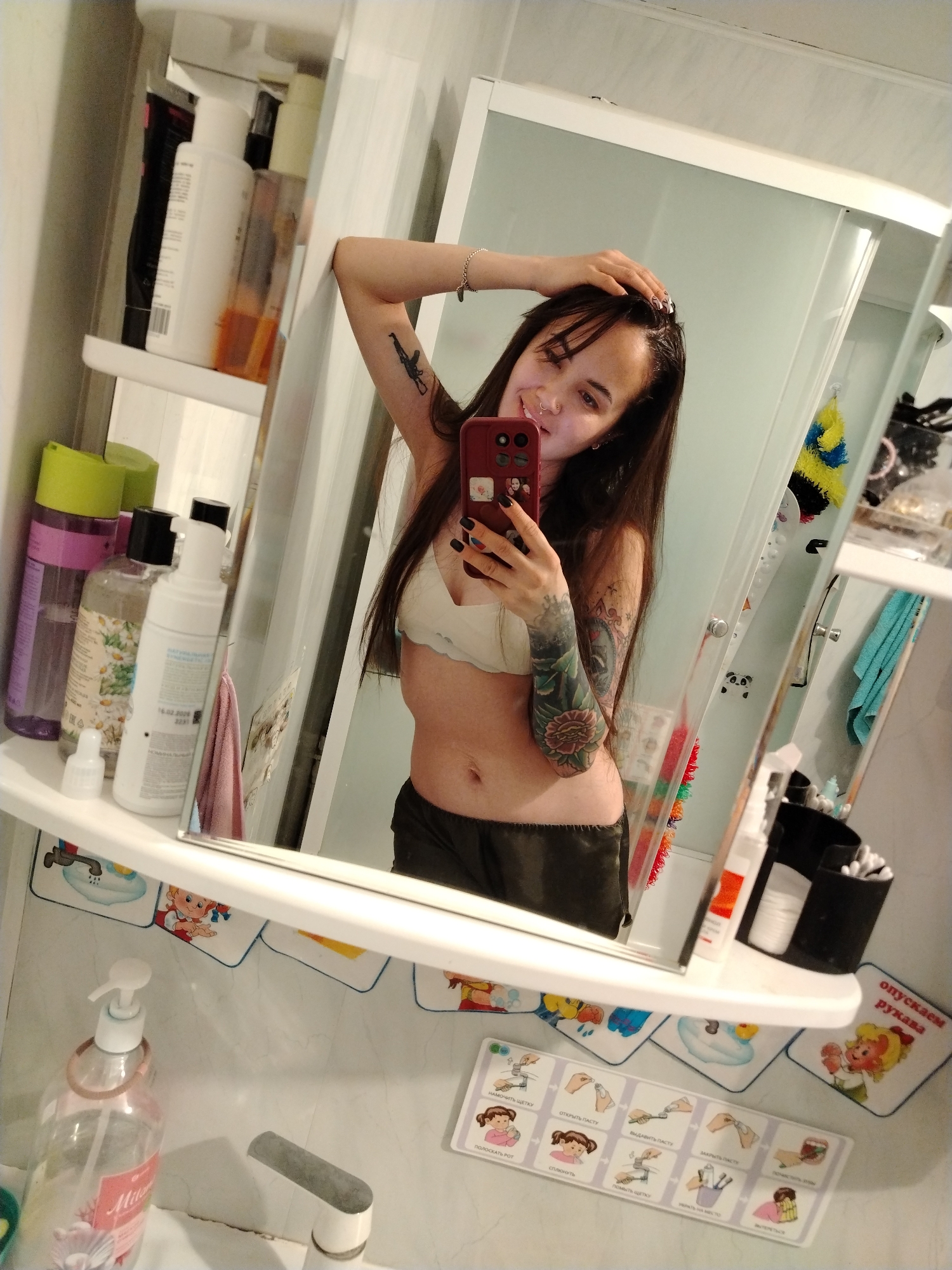 A woman is taking a selfie in a bathroom. She is holding a red smartphone in her right hand, her face partially obscured by the phone. She is wearing a bra and shorts. Her left arm is raised and touching her head. Both arms have tattoos. The bathroom has a mirror, sink, and shelves with various toiletries. The sink has a soap dispenser and faucet. A small, framed picture illustrating hand washing steps is visible below the mirror. The OCR text on the frame reads "16.02.2006", "НАМОЧИТЬ ЩЕТКУ", "ОТКРЫТЬ ПАСТУ", "ВЫДАВИТЬ ПАСТ", "ЗАКРЫТЬ ПАСТЕ", "ПОЛОСКАТЬ РОТ", "сплюнуть", and "опускаем рукава". The picture is located on the wall of a residential bathroom. No additional information about the location in Noviy, Russia is discernable.