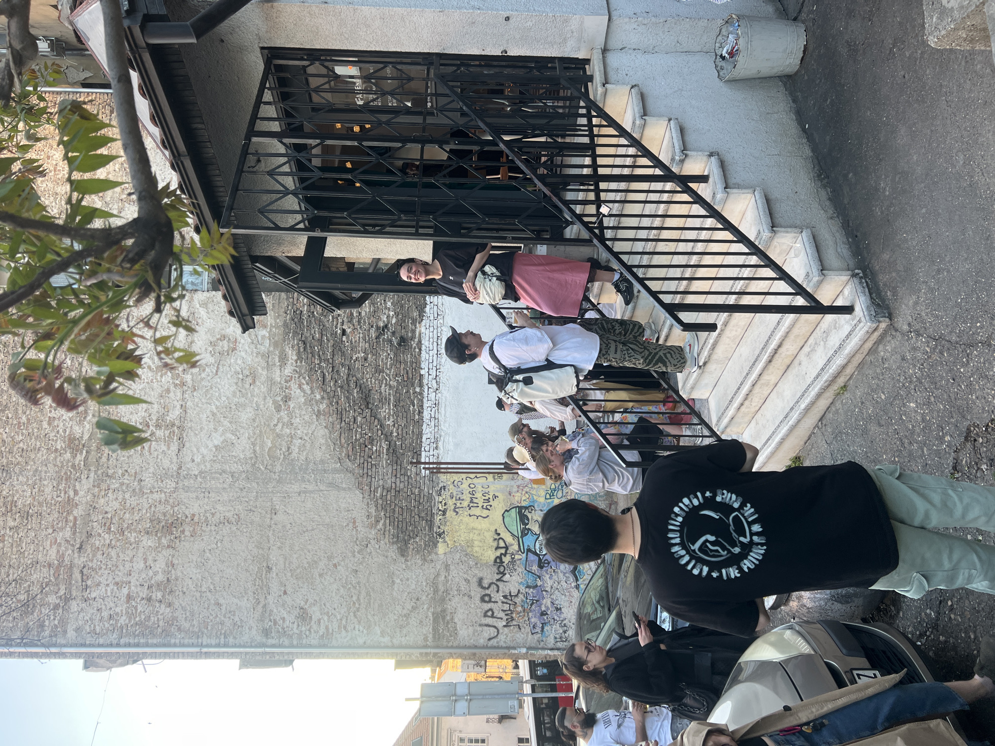 The image shows a group of people standing and sitting outside a building in Belgrade, Serbia. The building has a staircase leading up to an entrance with a black metal railing. Some people are standing on the staircase, while others are sitting or standing on the sidewalk. Graffiti is visible on the wall adjacent to the staircase. The setting appears to be an urban area with buildings and a street visible in the background. A tree with green leaves is visible in the upper left corner of the image.