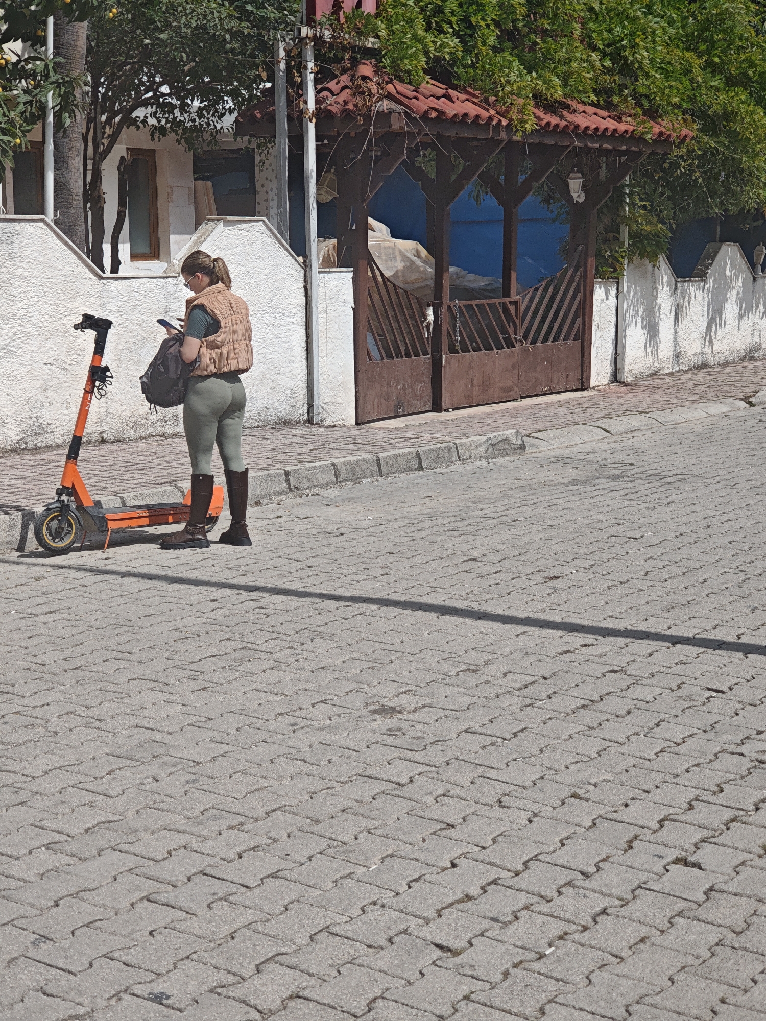 A woman stands on a cobblestone street next to an orange scooter in Kemer, Turkey. She wears a tan vest, olive leggings, and knee-high brown boots. She holds a cell phone in her hand and has a backpack on her shoulder. Behind her is a white stucco wall, a wooden gazebo-like structure, and a building with trees and plants. The street takes up much of the frame.