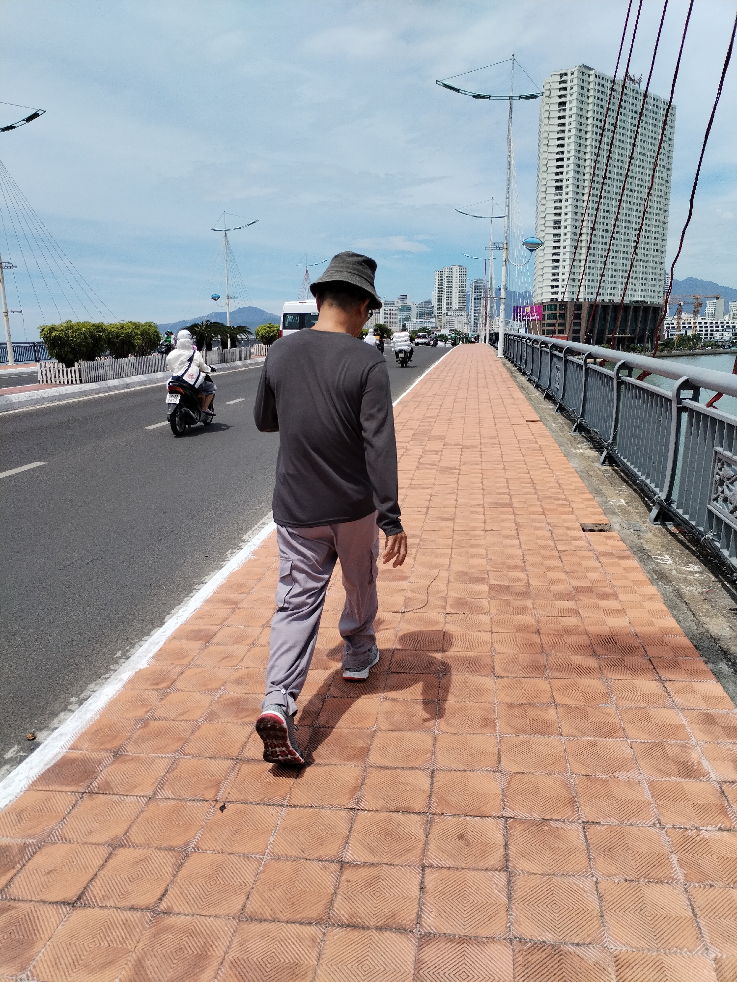 A man in a gray long-sleeved shirt, gray pants, and a bucket hat walks away from the camera on a pedestrian walkway paved with orange tiles. He is on a bridge in Nha Trang, Vietnam, with a multi-lane road to his left and a metal railing to his right overlooking a body of water. In the background, there are high-rise buildings, a large, white, modern skyscraper, and the cables of the bridge. Several motorcycles are visible on the road, and a white bus is further down the road. The sky is blue with scattered clouds. The scene appears to be during the daytime.