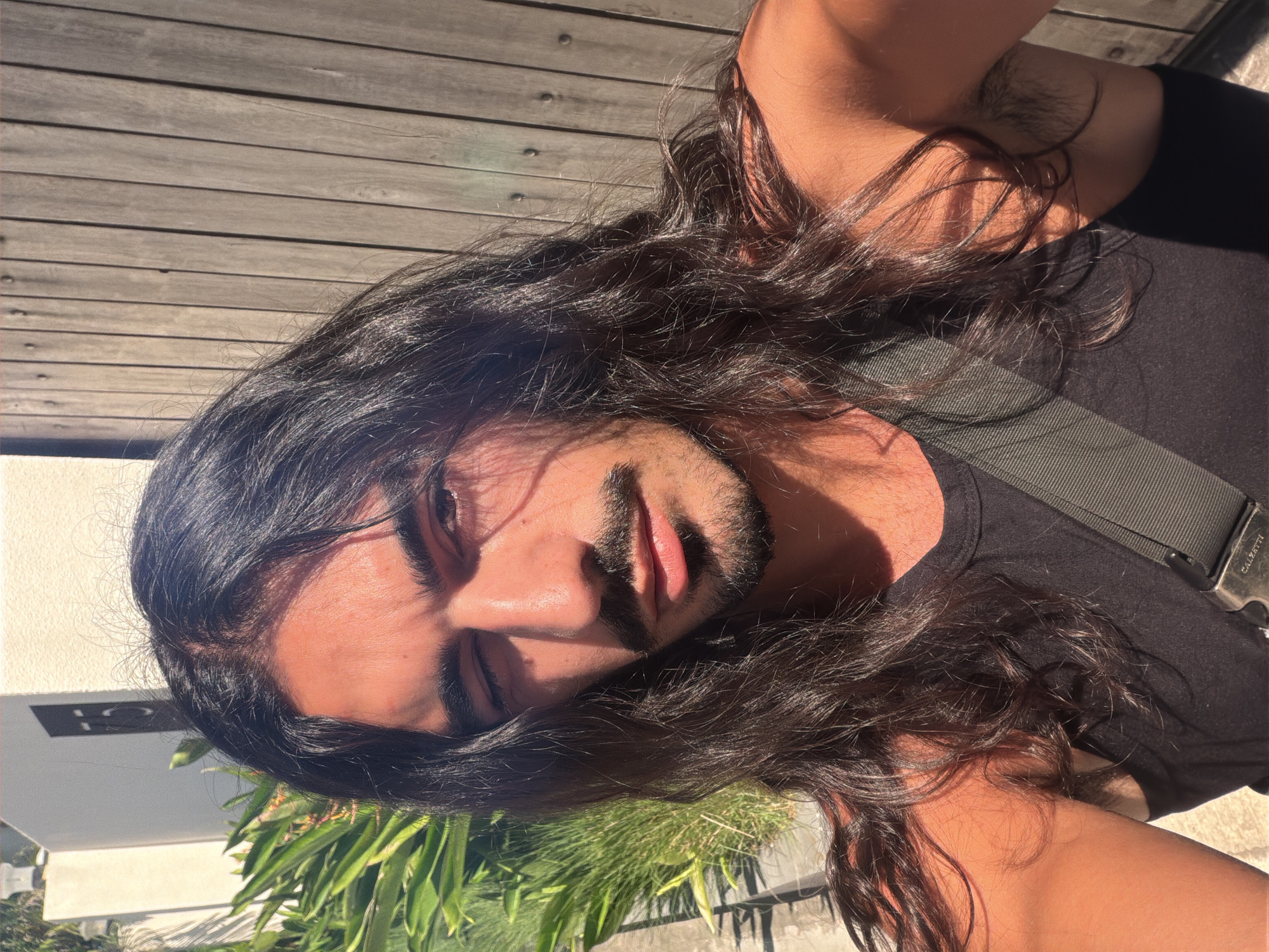 A medium shot depicts a man with long, dark brown hair, a beard, and a dark complexion, possibly of Indonesian heritage, wearing a black t-shirt. A green belt strap is visible across his chest. He is positioned outdoors during daylight, with a wooden wall or fence in the background and green foliage visible in the lower left corner. Given the prompt, this may be North Kuta, Indonesia.