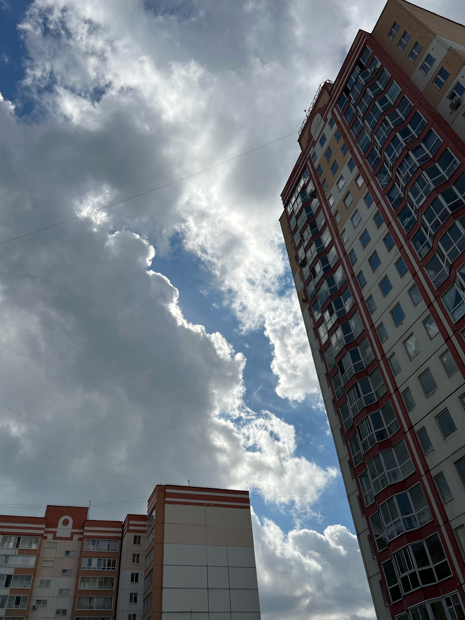 The image shows a view looking up at apartment buildings against a cloudy sky. In the foreground on the right is a tall, multi-story apartment building with a red and cream facade, featuring numerous windows, some with bay window designs. To the left, two shorter apartment buildings with similar architectural styles are visible. The sky is filled with a mix of white, fluffy cumulus clouds and darker, more ominous clouds, with patches of blue sky showing through. The perspective is from a low angle, emphasizing the height of the buildings and the expanse of the sky. The overall scene suggests an urban environment with residential architecture under a dynamic sky. The location is identified as Tomsk, Russia.