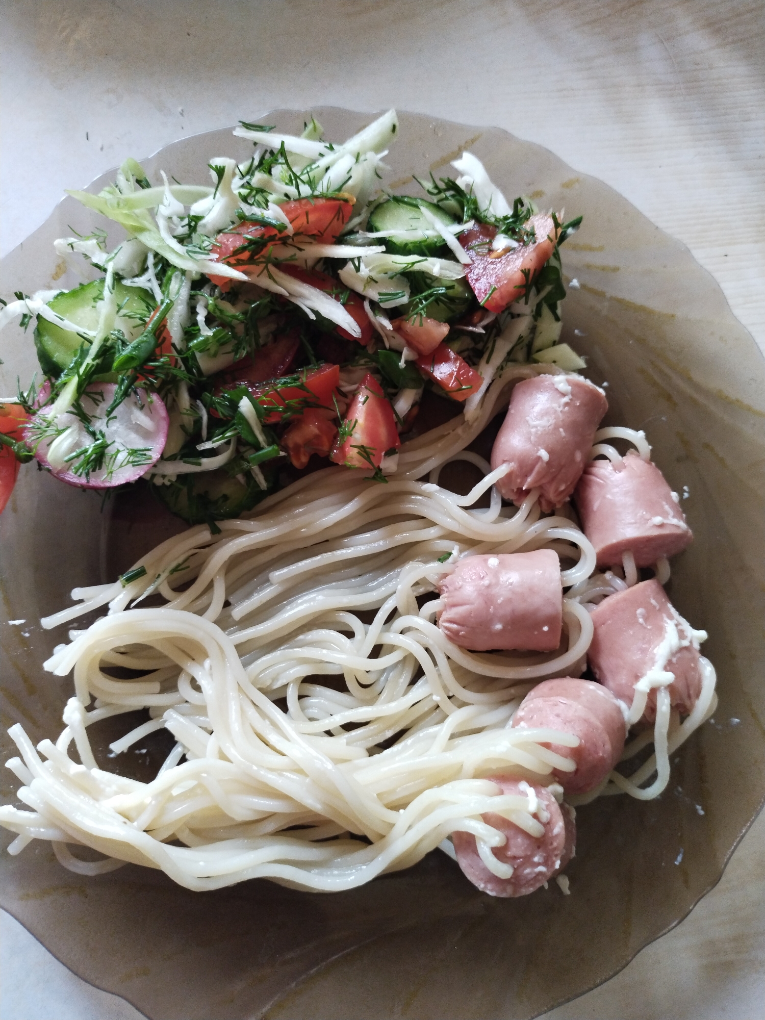 A close-up, overhead photograph depicts a meal served in a shallow, irregularly shaped dish of light brown or beige material. The dish contains three primary components: a fresh salad, cooked spaghetti, and five small sausages. The salad, positioned in the upper portion of the dish, consists of shredded white cabbage, diced red tomatoes, sliced green cucumbers, round slices of pinkish-white radishes, and finely chopped green dill, all appearing lightly dressed. Below the salad and extending to the bottom of the dish are strands of light beige spaghetti or vermicelli, some of which are entwined with a white, creamy substance. Embedded within the spaghetti are five cylindrical pieces of light pinkish-brown sausage, each approximately 2-3 cm in length. Several spaghetti strands are visibly threaded through the cut sections of the sausages. The same white, creamy substance present on the spaghetti is also visible adhering to the surfaces of the sausages. The background features a light, out-of-focus surface, possibly wood or a countertop. The scene is located in Horishni Plavni, Ukraine. No individuals are visible.