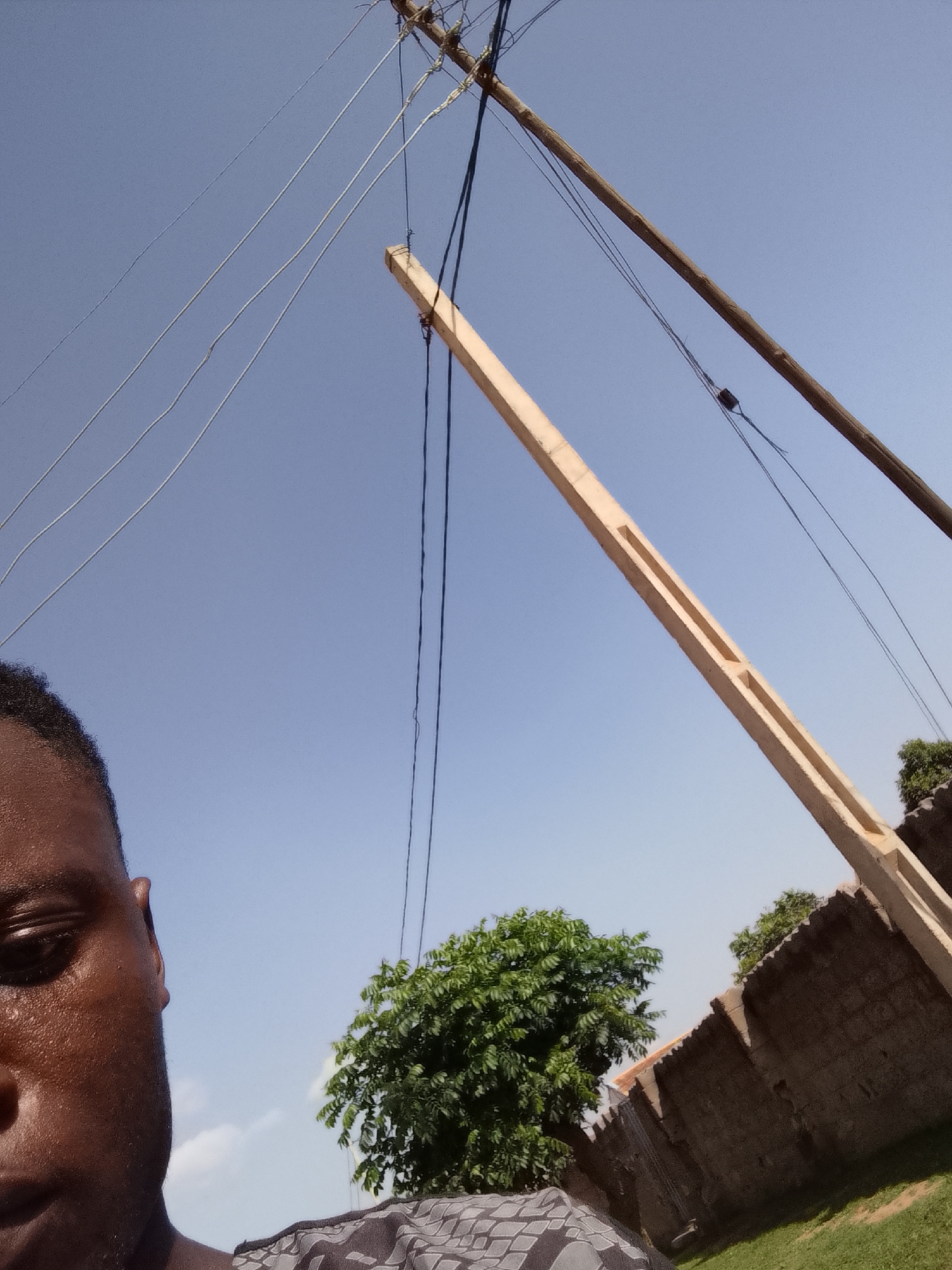 In Wauro Leggal, Nigeria, this image captures a person's face and shoulder in the foreground, looking up towards a clear blue sky. Overhead, a power line with multiple wires stretches across the frame, supported by a wooden pole and a concrete pole. In the mid-ground, a dense green tree stands beside a corrugated metal fence. The background is dominated by the sky, with hints of clouds visible. The lighting suggests it is daytime.