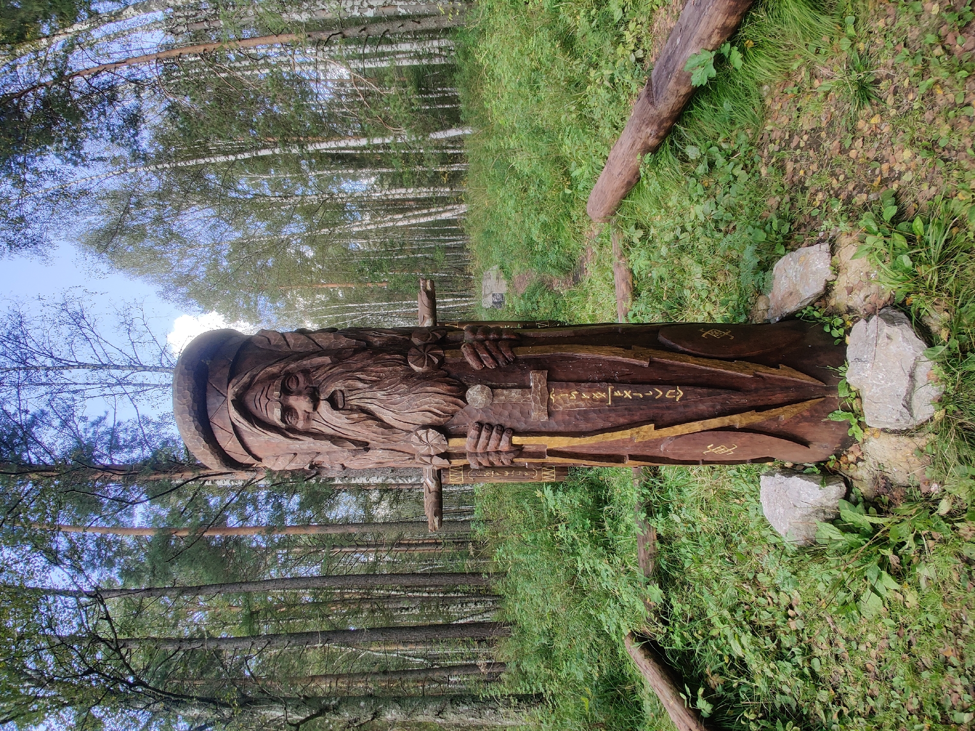 A wooden sculpture of a bearded figure, possibly a Slavic deity or spirit, stands prominently in a forest clearing. The sculpture is carved from a large tree trunk and depicts a weathered face with deep-set eyes, a long beard, and a headdress. The figure holds what appears to be a sword or staff. The sculpture is decorated with yellow paint and runes or symbols. It is supported by stones at its base. The surrounding environment is a forest with tall trees, including pines and birches, and dense undergrowth of green plants and grass. Fallen logs are visible on the ground in front of and to the sides of the sculpture. The sky is visible through the trees, with some clouds. The location is identified as the city of Bazhukovo, Russia.