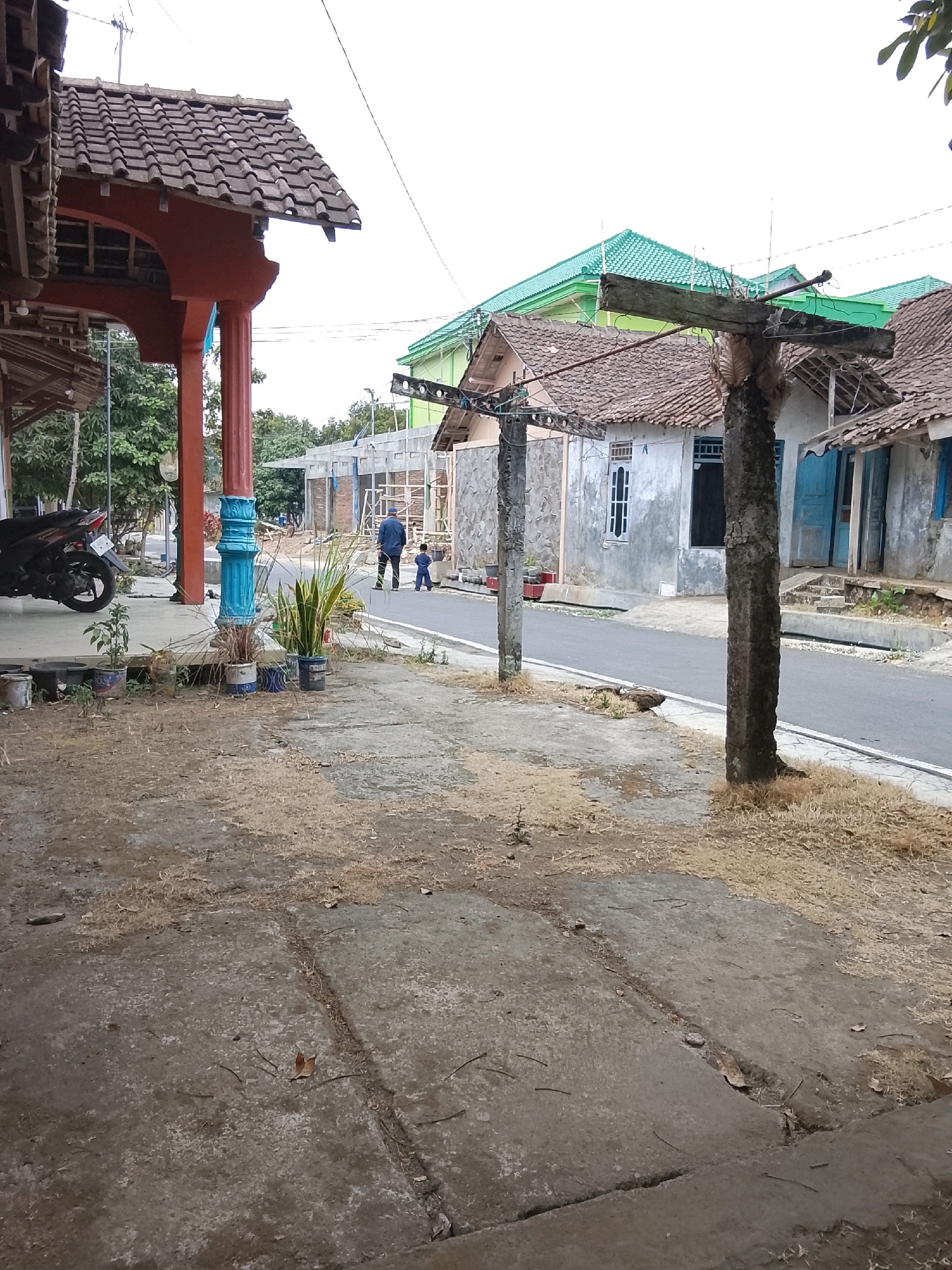 The image depicts a street scene in Salatiga, Indonesia. The foreground shows a concrete pathway with dry grass and some fallen leaves. A motorcycle is parked on the left, under a covered area with a tiled roof supported by an orange pillar with blue decorative elements. Beyond this, a paved road runs through a residential area. On the right side of the road, houses with tiled roofs and light-colored walls are visible. One house has blue doors and steps leading up to it. Further down the street, construction is underway with visible scaffolding and partially built structures. In the middle ground, a man in a blue shirt and dark pants is walking away from the camera, with a young child in a blue outfit walking beside him. The sky is overcast. The overall environment suggests a modest, lived-in neighborhood undergoing some development.